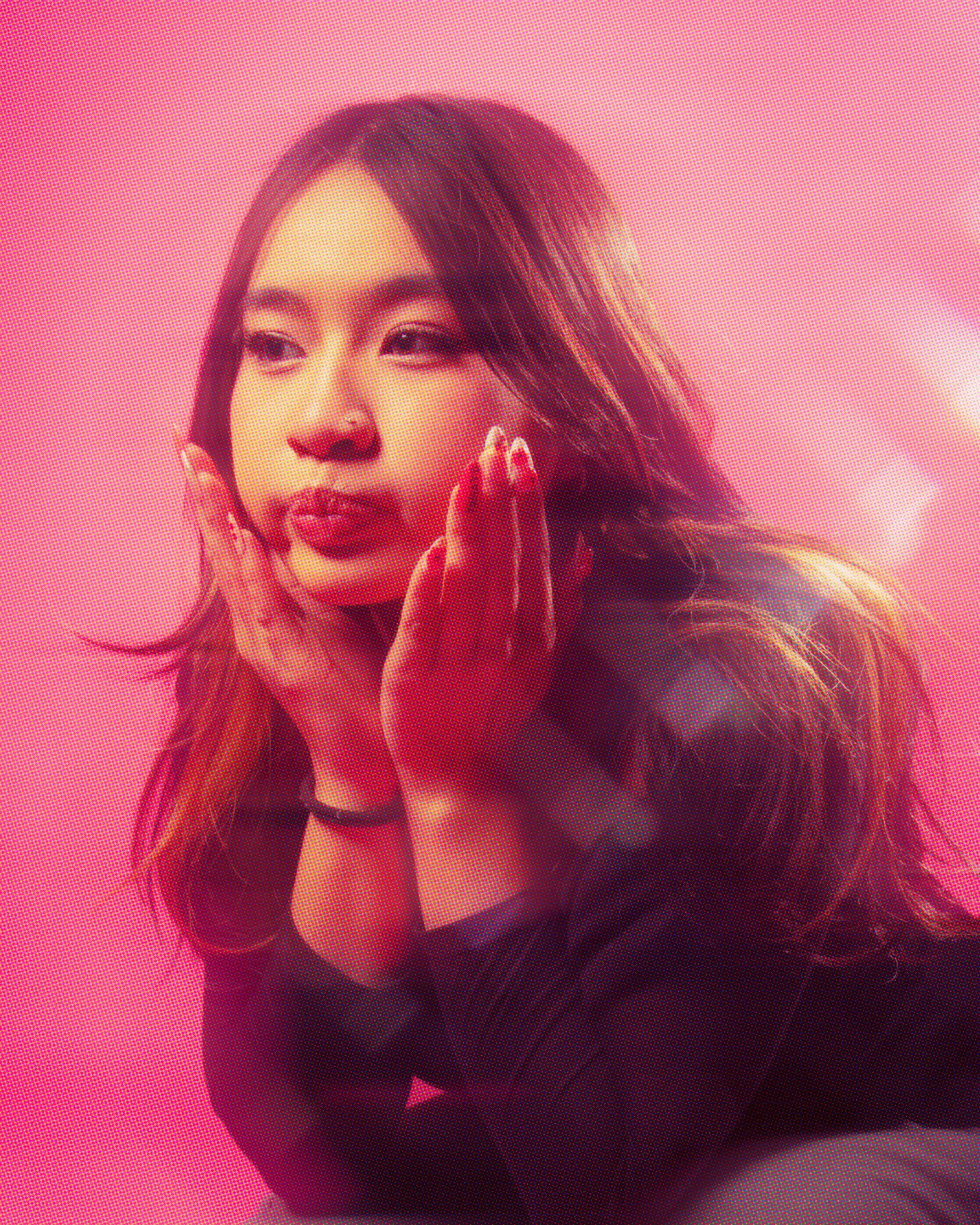 A woman with long, wavy hair and makeup, holding her face with both hands, in front of a pink background.