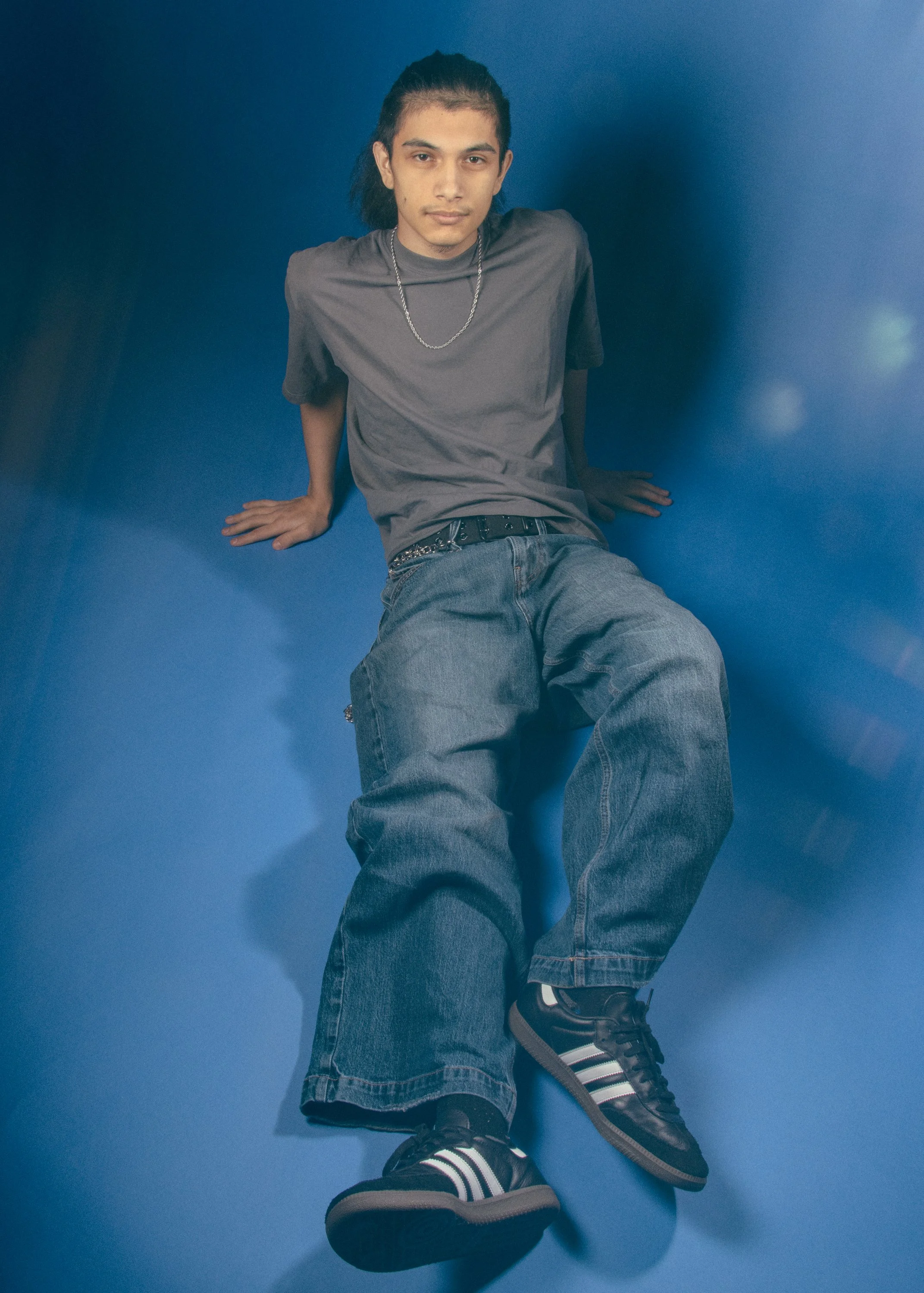 A young man with long dark hair in a ponytail, wearing a gray t-shirt, baggy jeans, black sneakers with white stripes, sitting against a blue background.