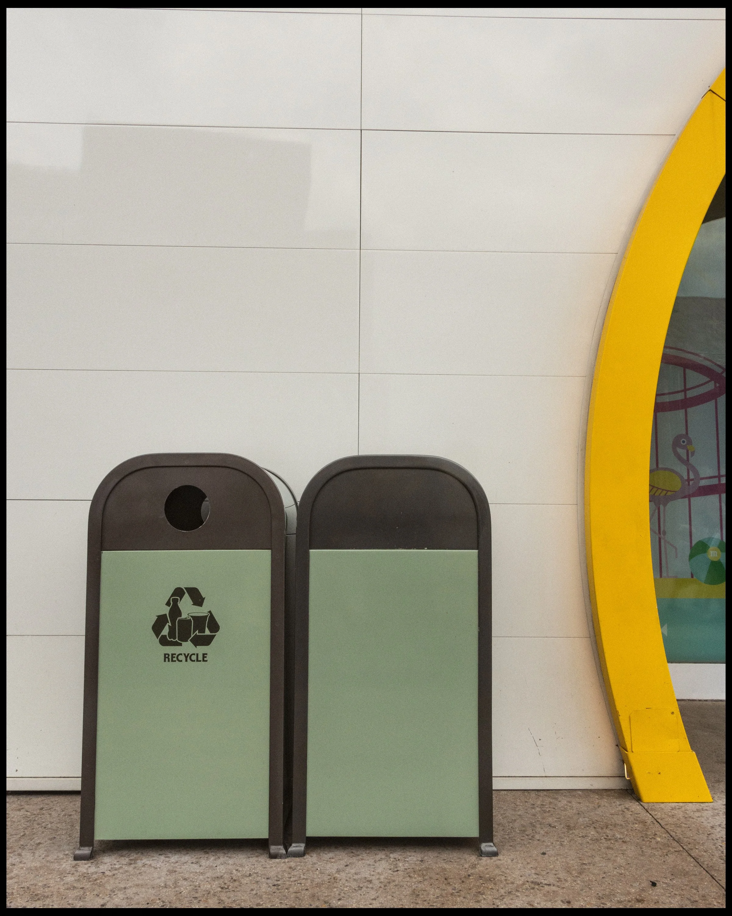 A line of gray chairs with rounded backs, positioned in front of a white tiled wall. A green recycling bin with a black recycling symbol is in front of the chairs. On the right side, a yellow safety barrier curves around a partial glass wall with col