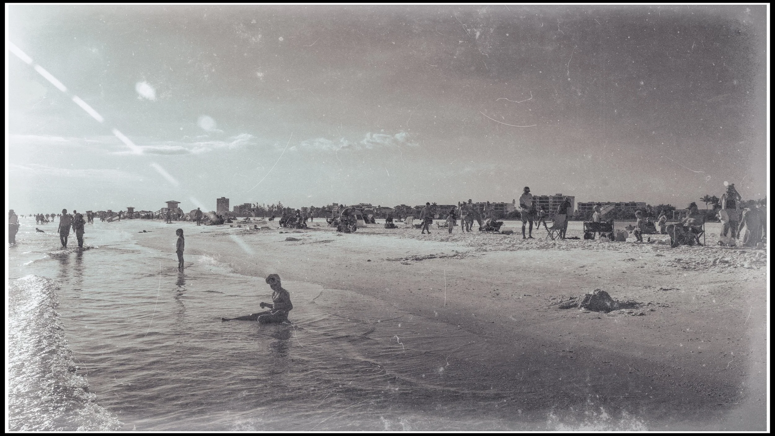 Black and white photograph of a crowded beach with people walking, sitting, and engaging in activities, with buildings in the background and the ocean in the foreground.