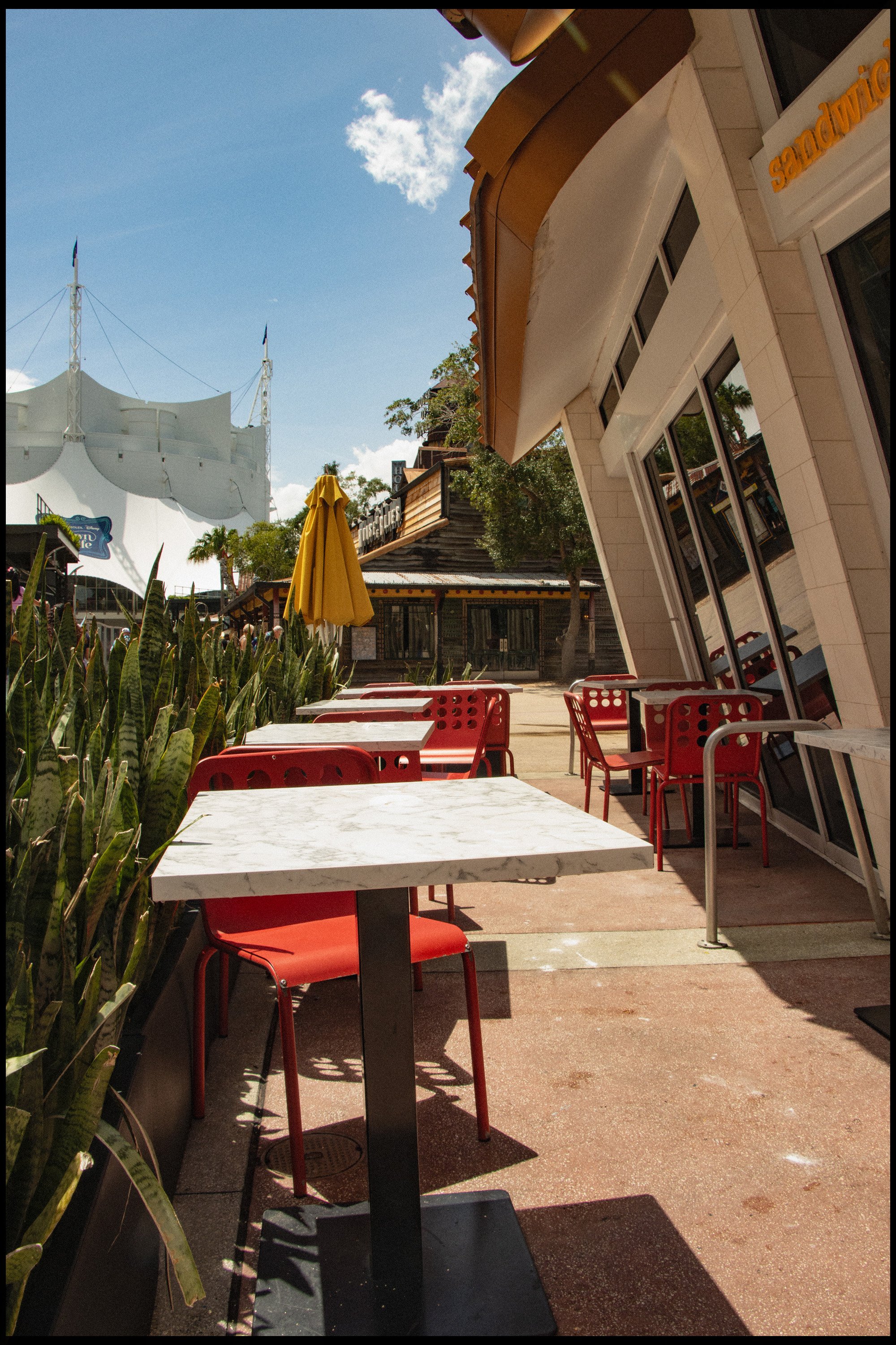 Empty outdoor patio seating area with red chairs and white marble tables, yellow umbrella, and surrounding plants, outside a modern building with large windows and signage, under a partly cloudy blue sky.