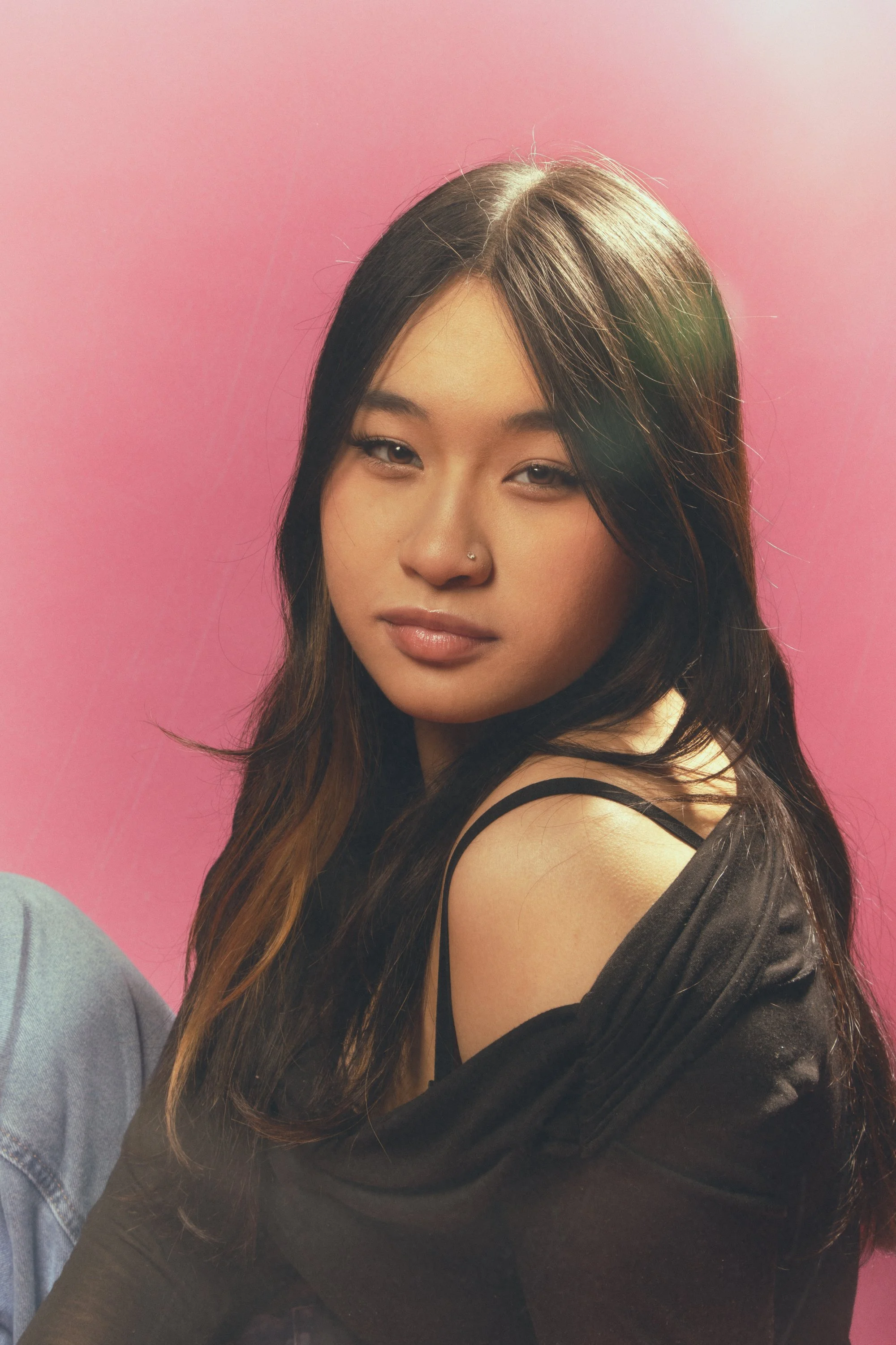 A woman with long dark hair and a nose piercing, wearing a black off-shoulder top, sitting against a pink background
