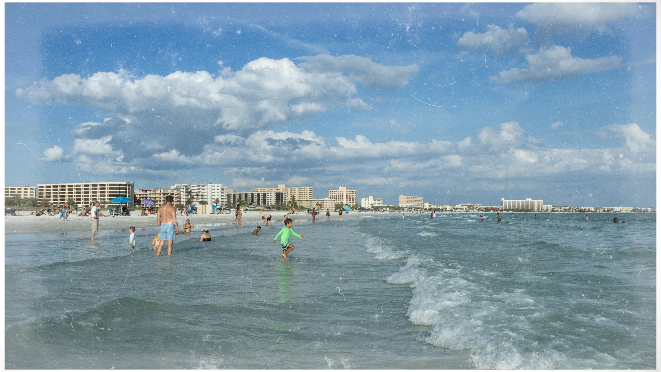 People swimming and relaxing on a crowded beach with high-rise buildings in the background under a partly cloudy sky.