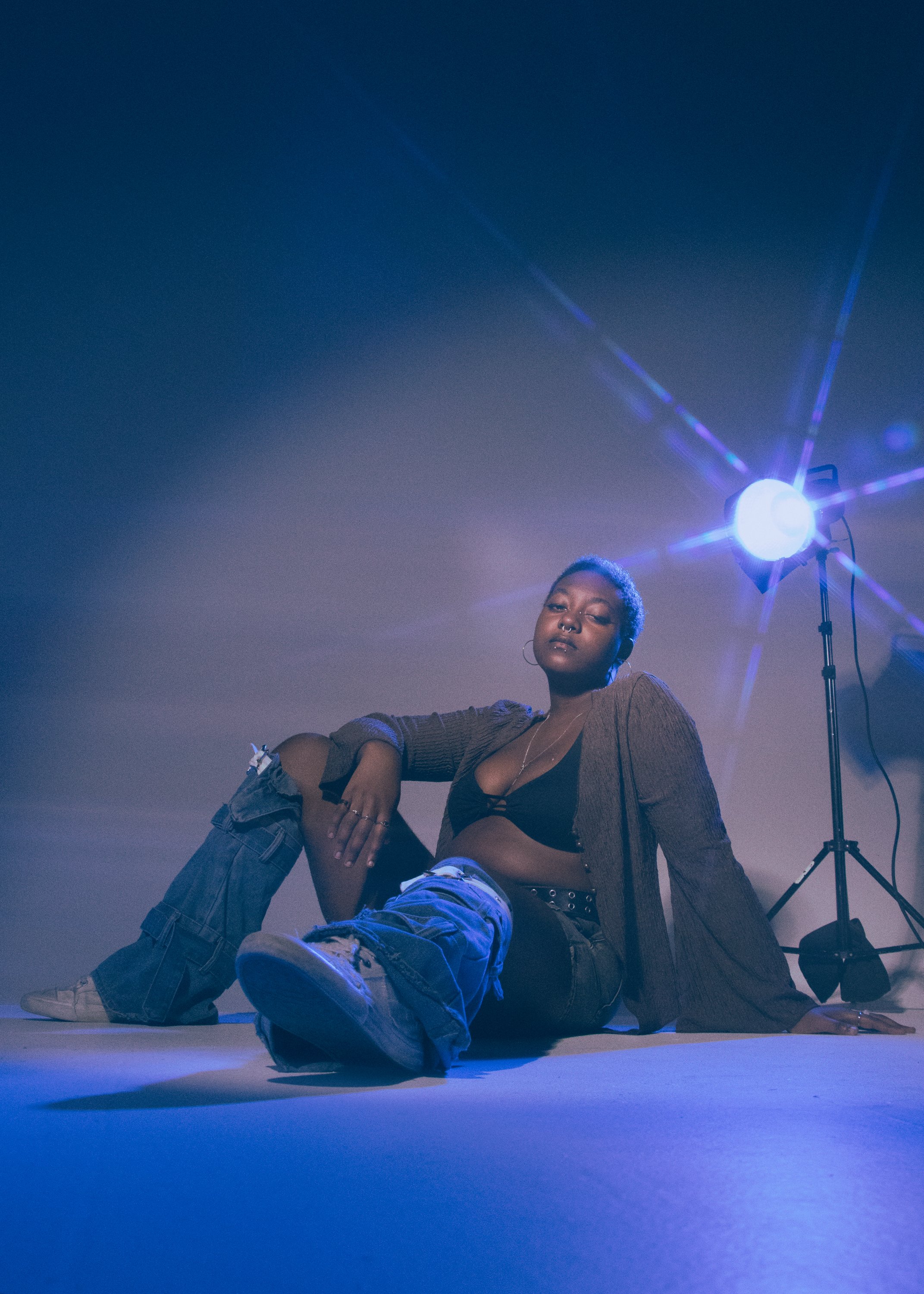 A woman with short hair and piercings poses on the floor with a man lying down, surrounded by blue and purple lighting, with a bright light and stand in the background.
