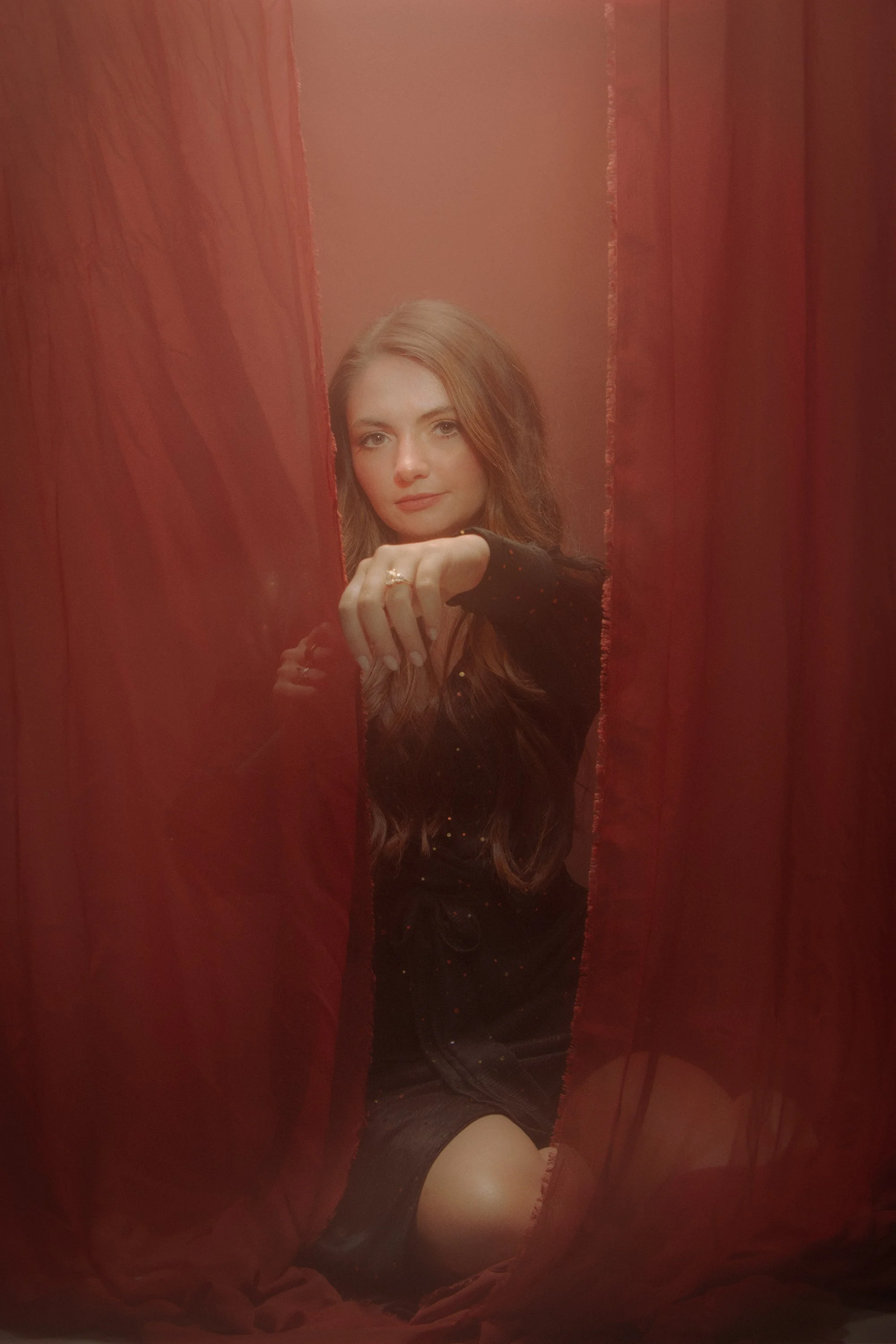 A woman taking a mirror selfie behind red curtains. She has long, wavy hair and is wearing a black dress and thigh-high stockings.