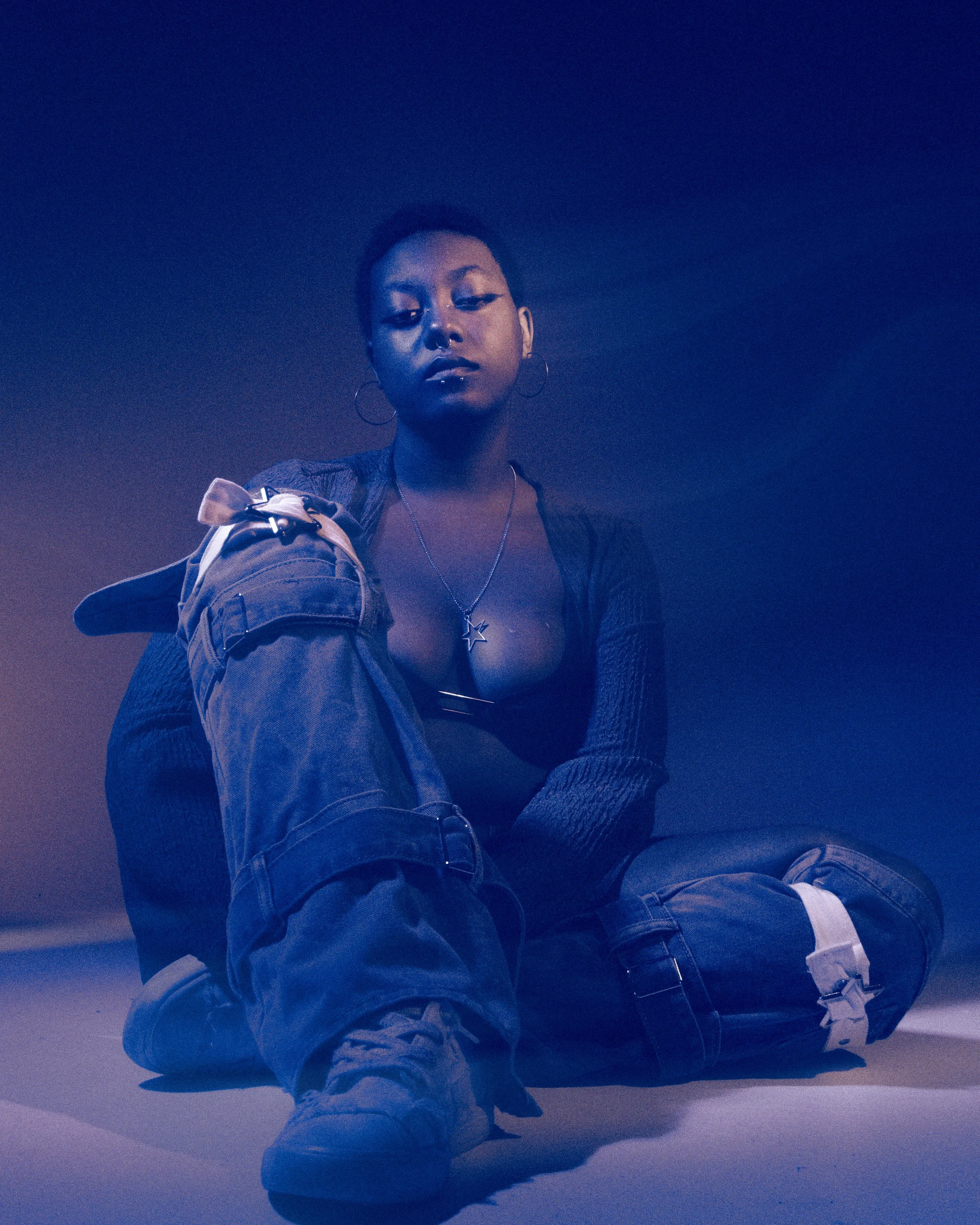 A woman with short hair, wearing hoop earrings, a star necklace, and a black jacket, sitting on the floor against a dark background, with blue and purple lighting.