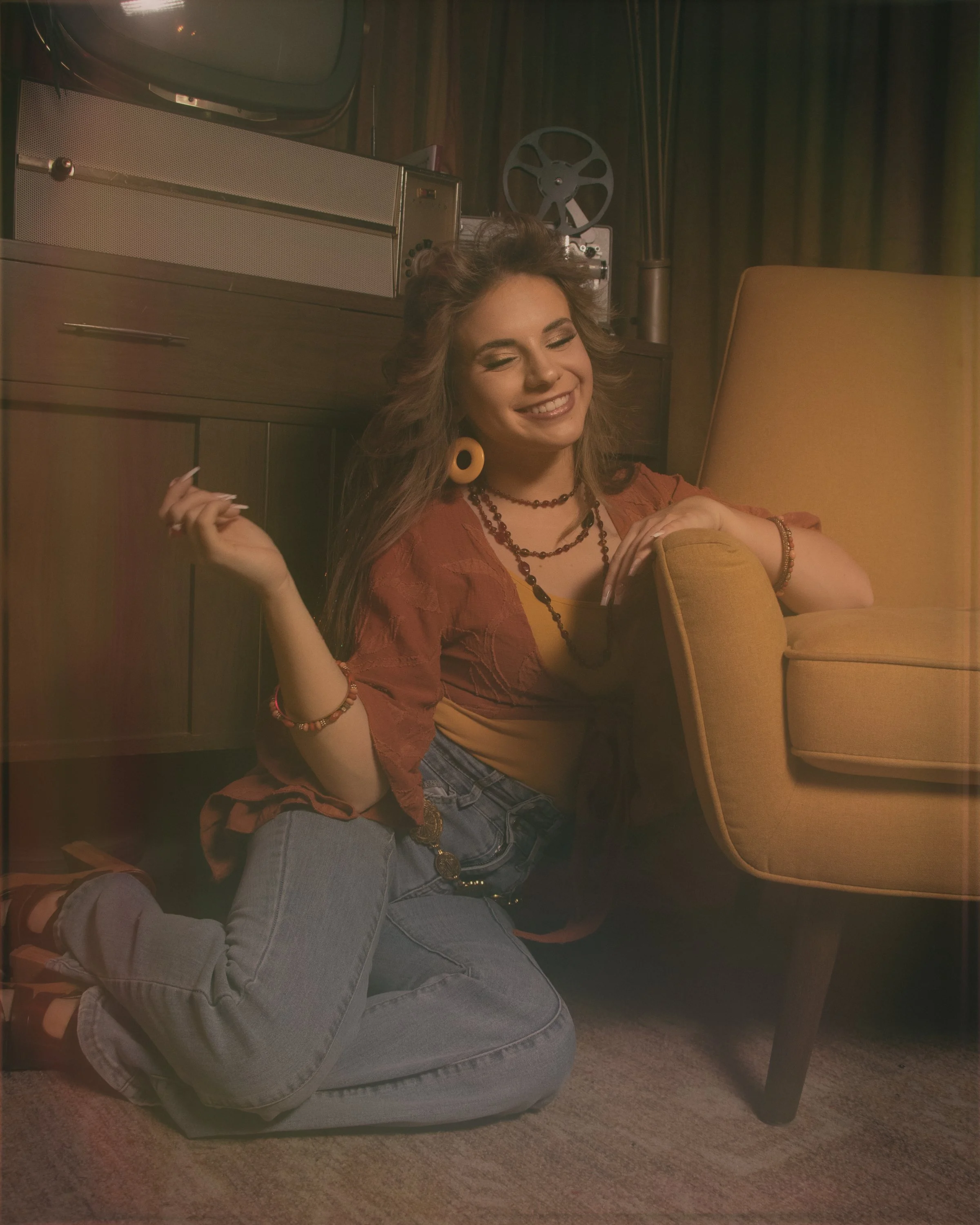 A young woman with curly hair, wearing earrings and layered necklaces, is sitting on the floor next to a mid-century modern lounge chair. She has a cheerful expression with eyes closed and a genuine smile. She is dressed in a yellow top with a reddis