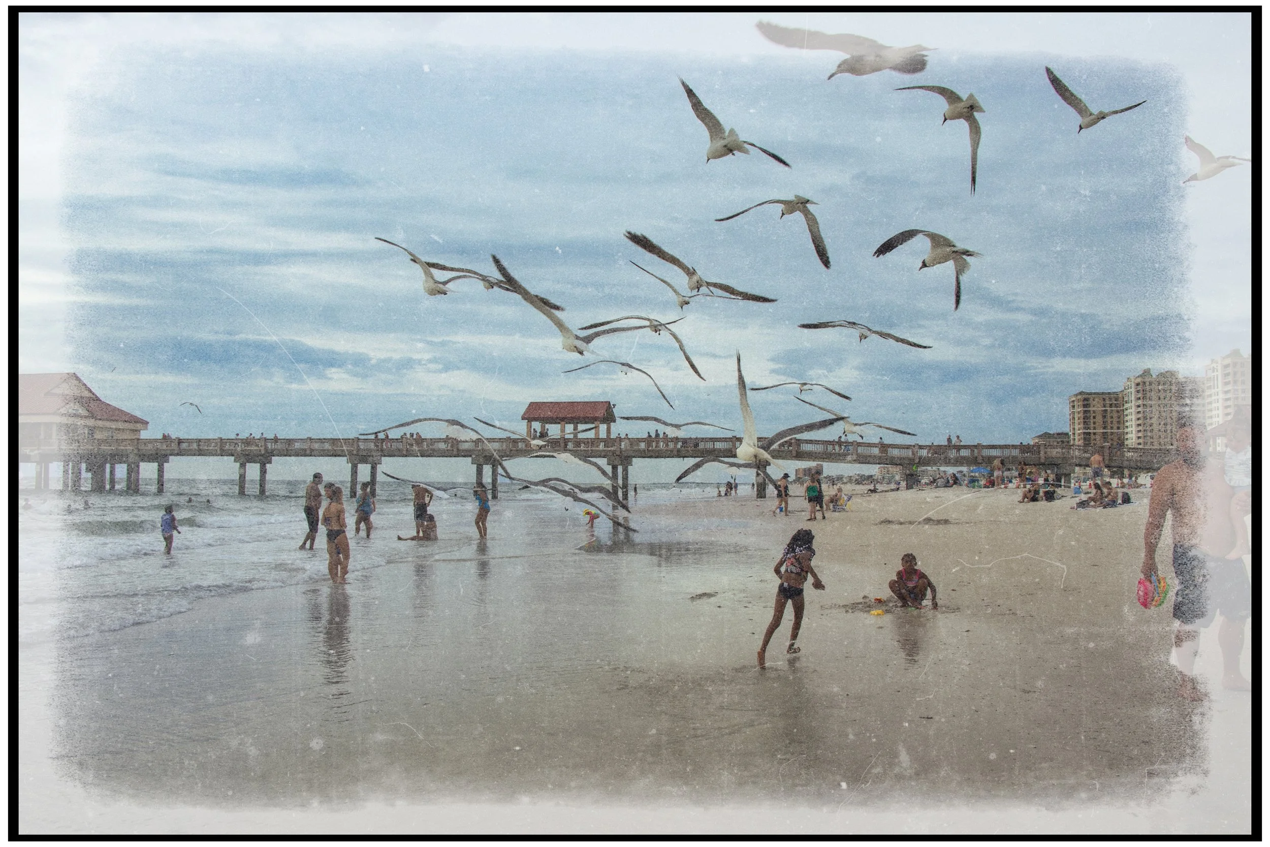 Seagulls flying over a crowded beach with people swimming and playing in the water, and a pier with a gazebo in the background.