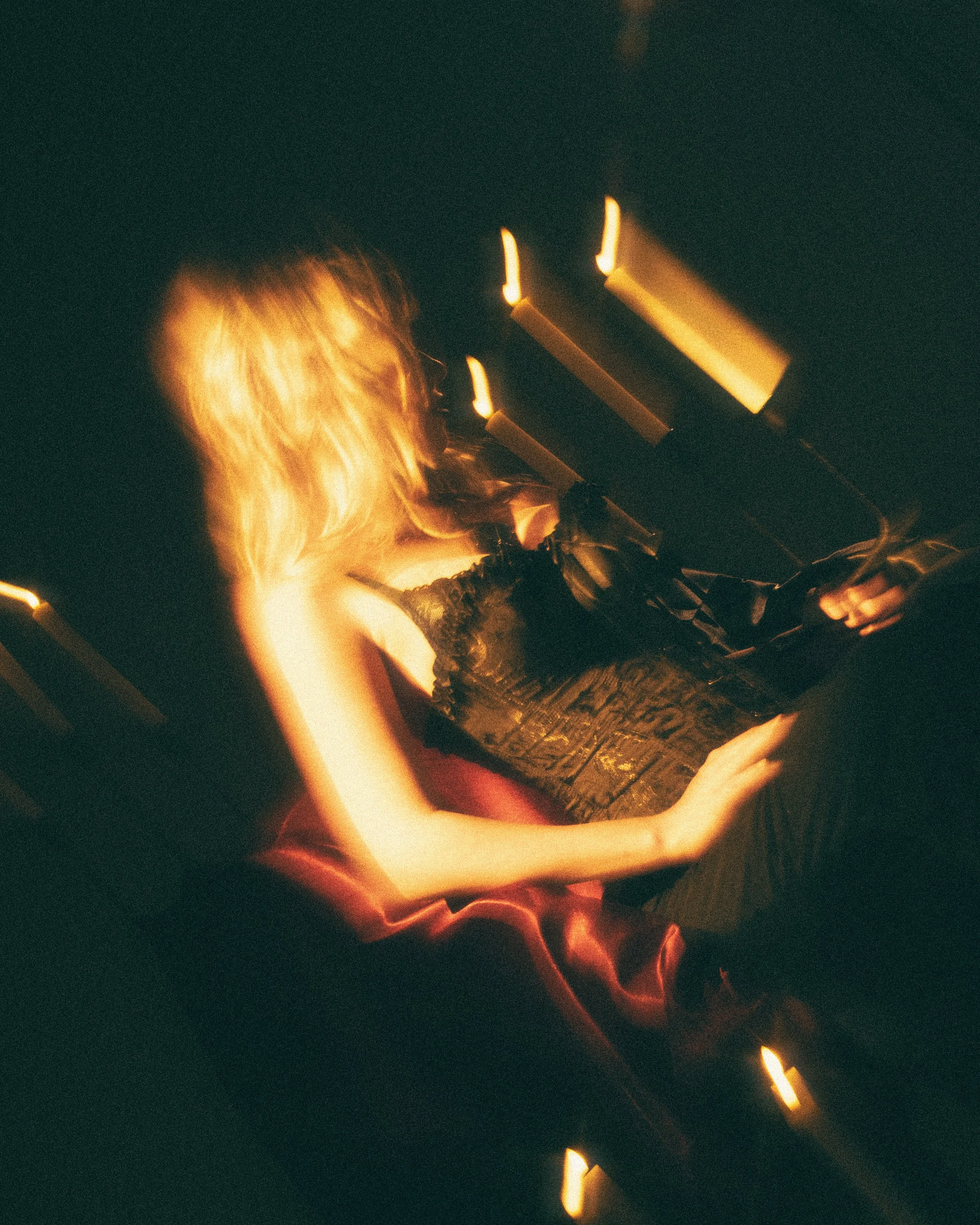 A woman with blonde hair sitting in a dark room, illuminated by several candles placed at different heights around her. The candles cast a warm glow on her face and surroundings.