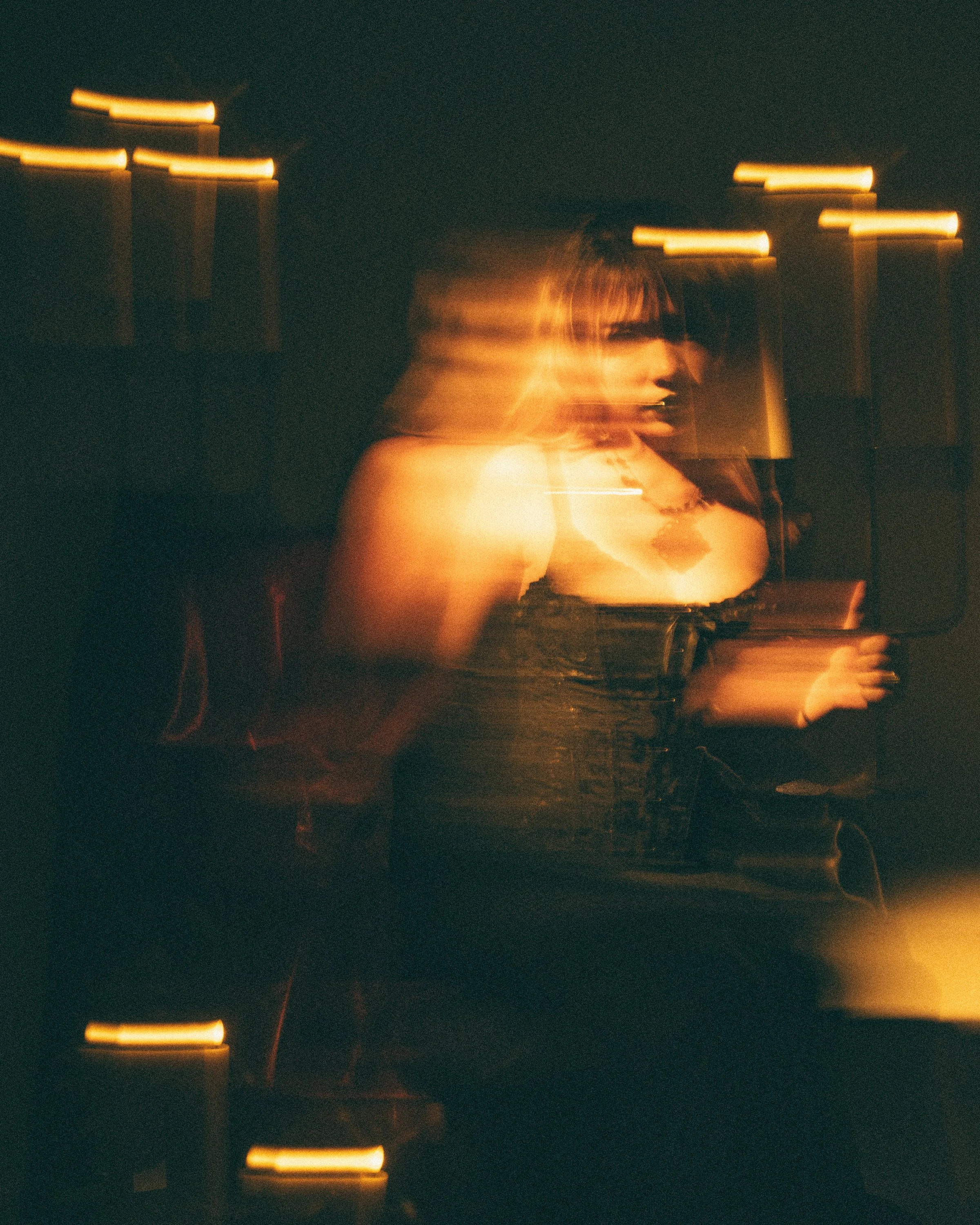 A woman with brown hair in a black top, captured with motion blur at night with city lights in the background.