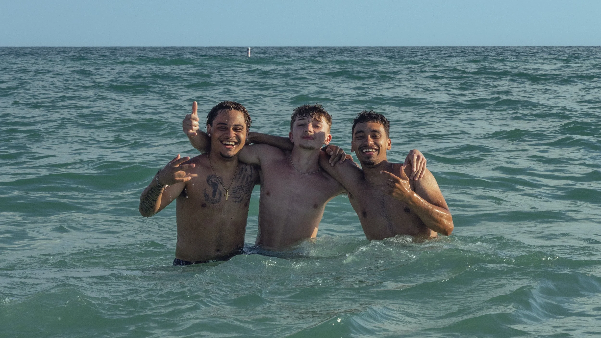 Three young men are enjoying their time in the ocean, smiling and posing for the photo with their arms around each other, surrounded by clear water and a calm sky.
