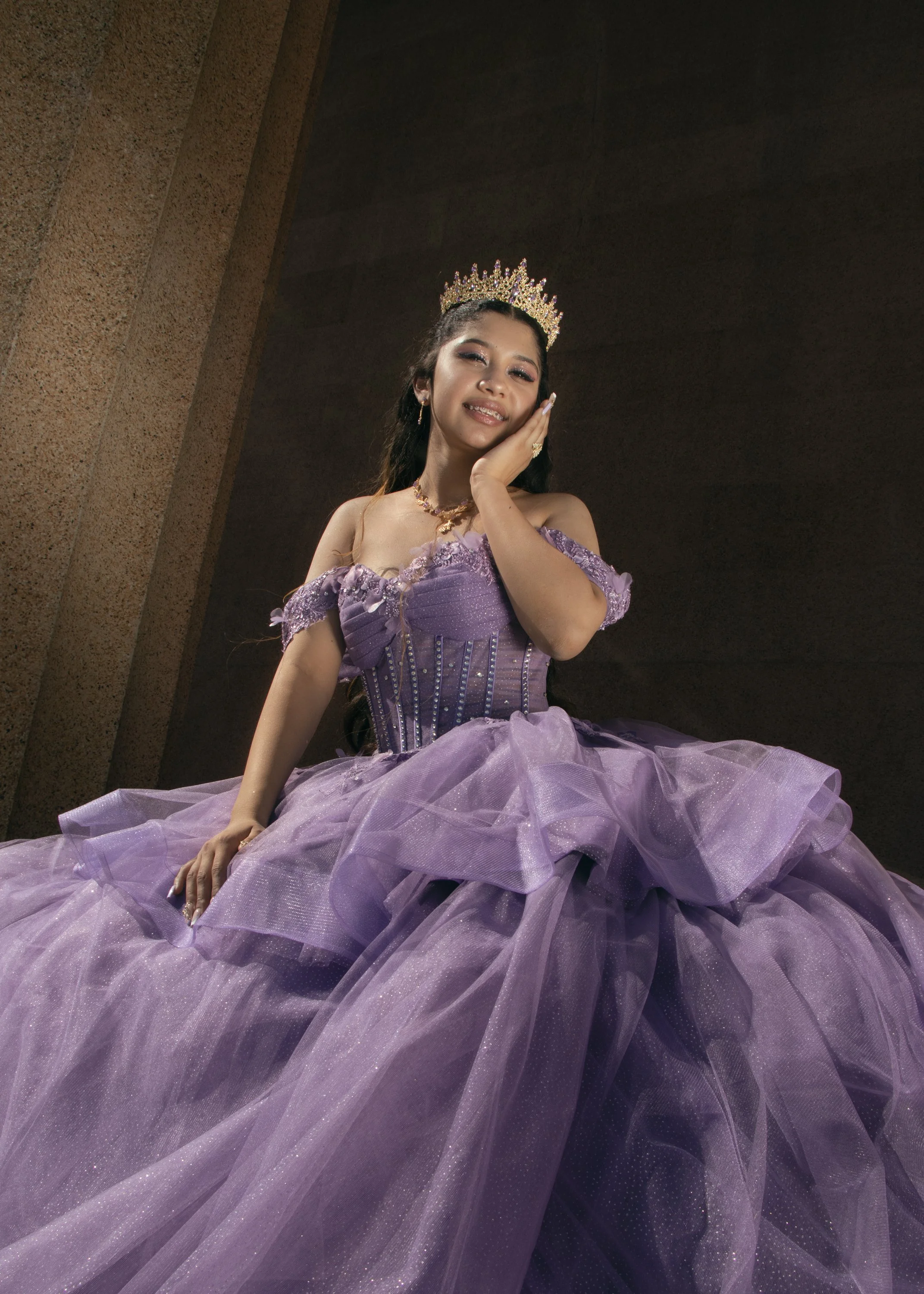 Young woman wearing a purple gown with a tiara, posing indoors against a textured wall.