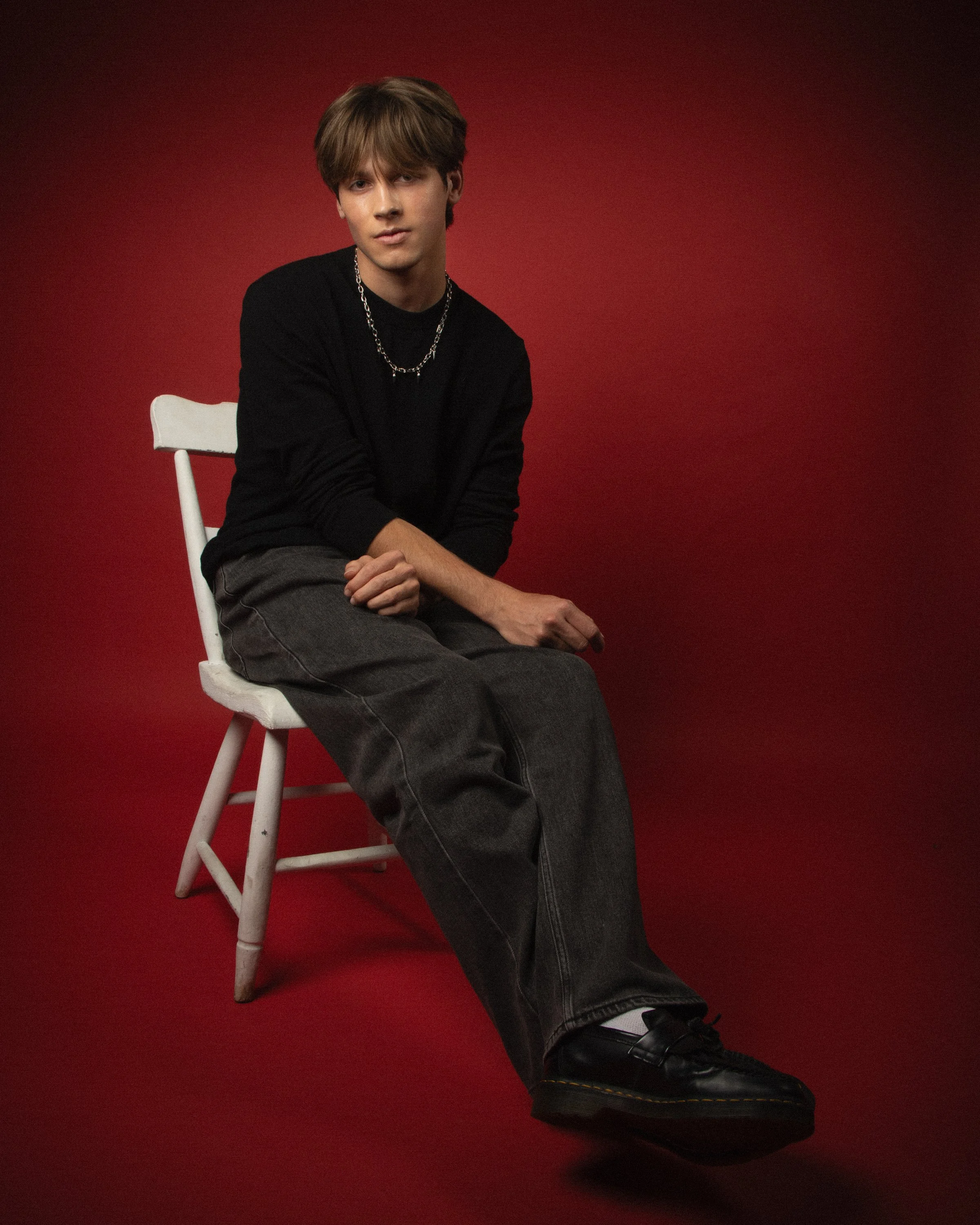 A young man with brown hair sitting casually on a white chair against a red background. He is wearing a black shirt, a silver chain necklace, gray pants, and black shoes. His right arm rests on his left knee, and he is looking directly at the camera.