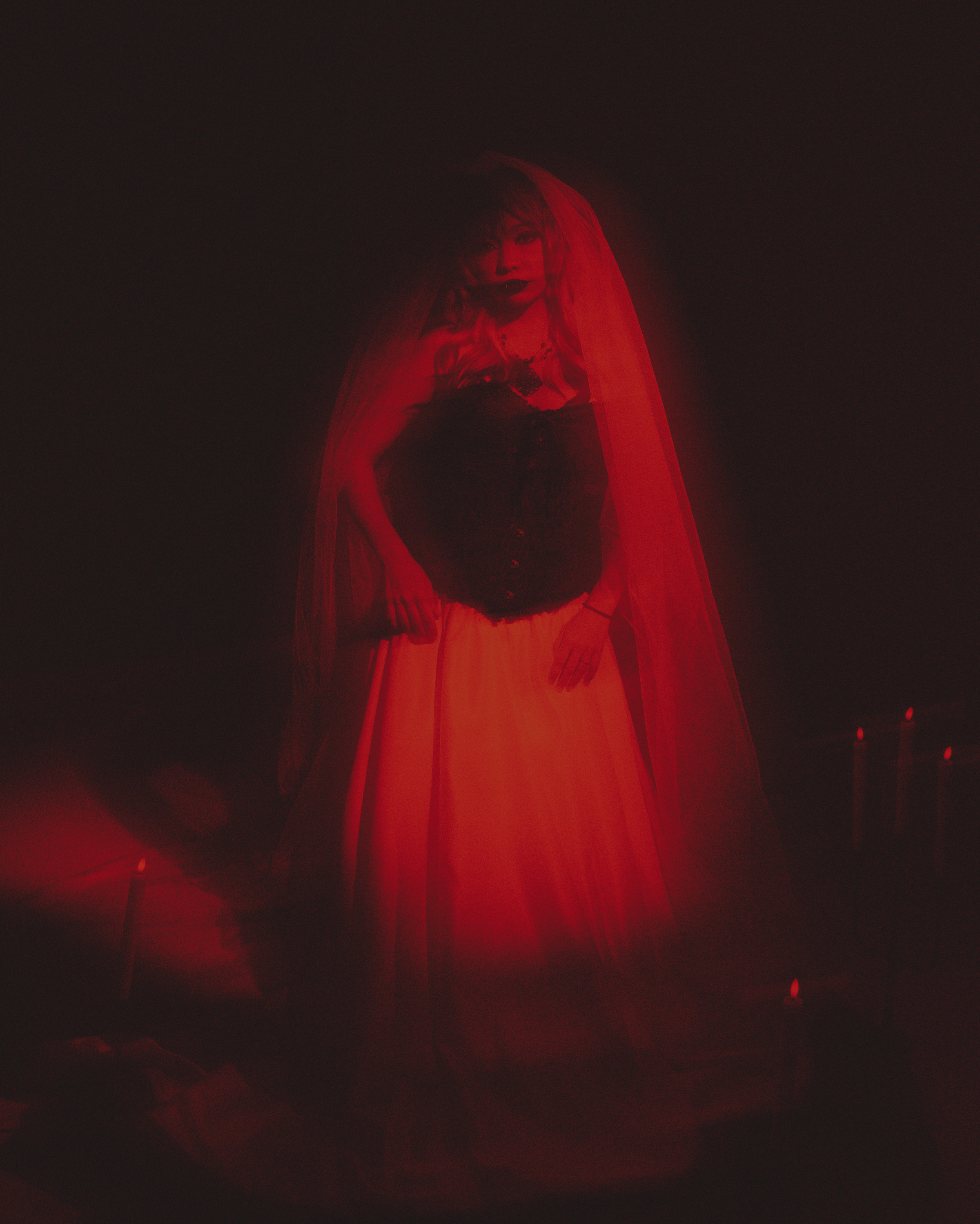 A woman dressed in black, wearing a red veil, standing in a dark setting with dim red lighting.