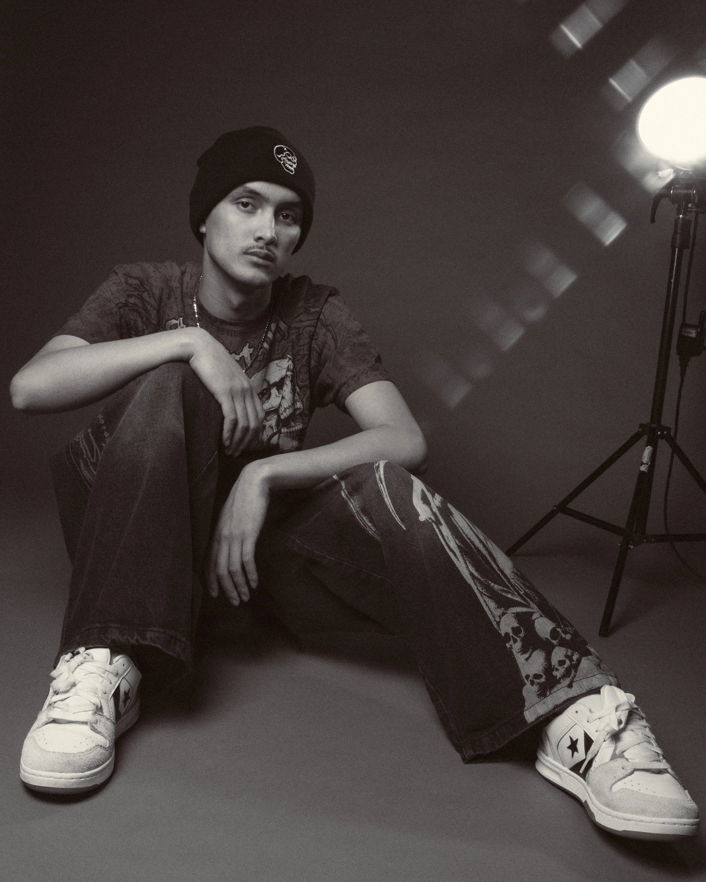 Young man sitting on the floor, wearing a black beanie, graphic t-shirt, baggy jeans, and white sneakers with black star logo, in a photography studio with studio light on a tripod to the right.