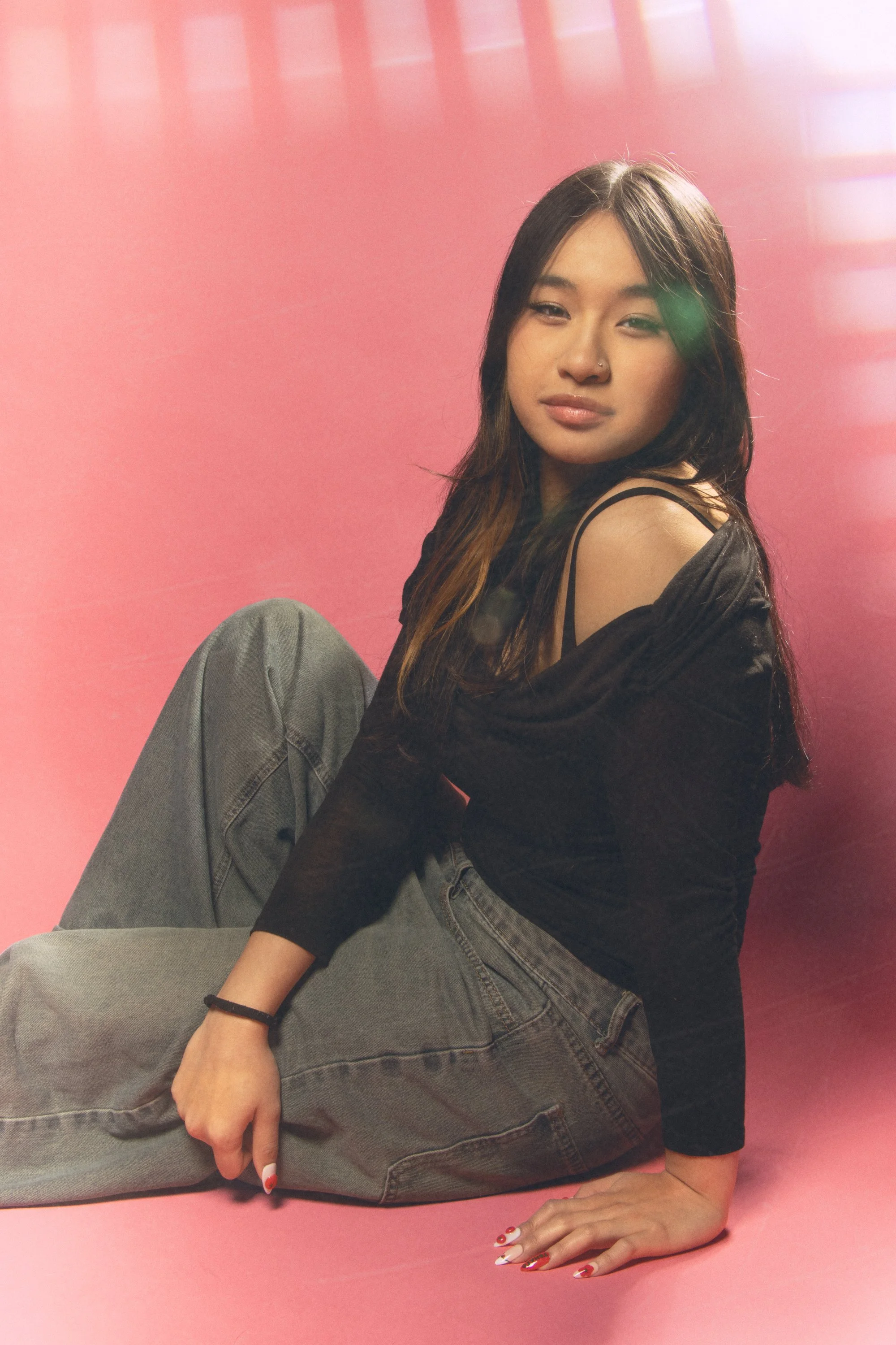 A young woman with dark hair and a nose piercing poses against a pink background. She is wearing a black off-shoulder top with spaghetti straps and gray jeans, sitting with one arm on the ground and the other resting on her knee, showing her painted 