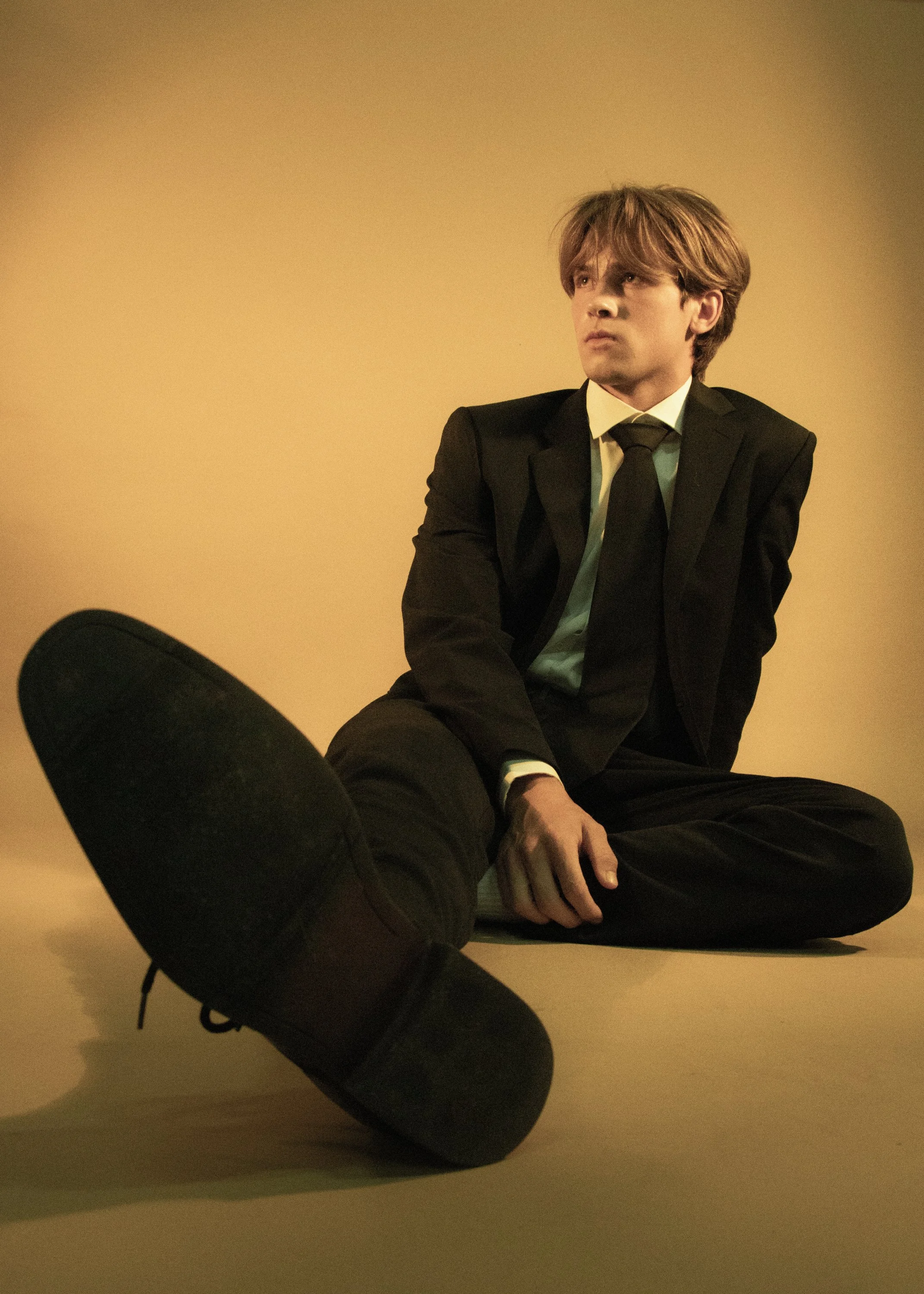 A person dressed in a black suit and tie sitting against a plain beige wall, with another person's leg and foot in the foreground.