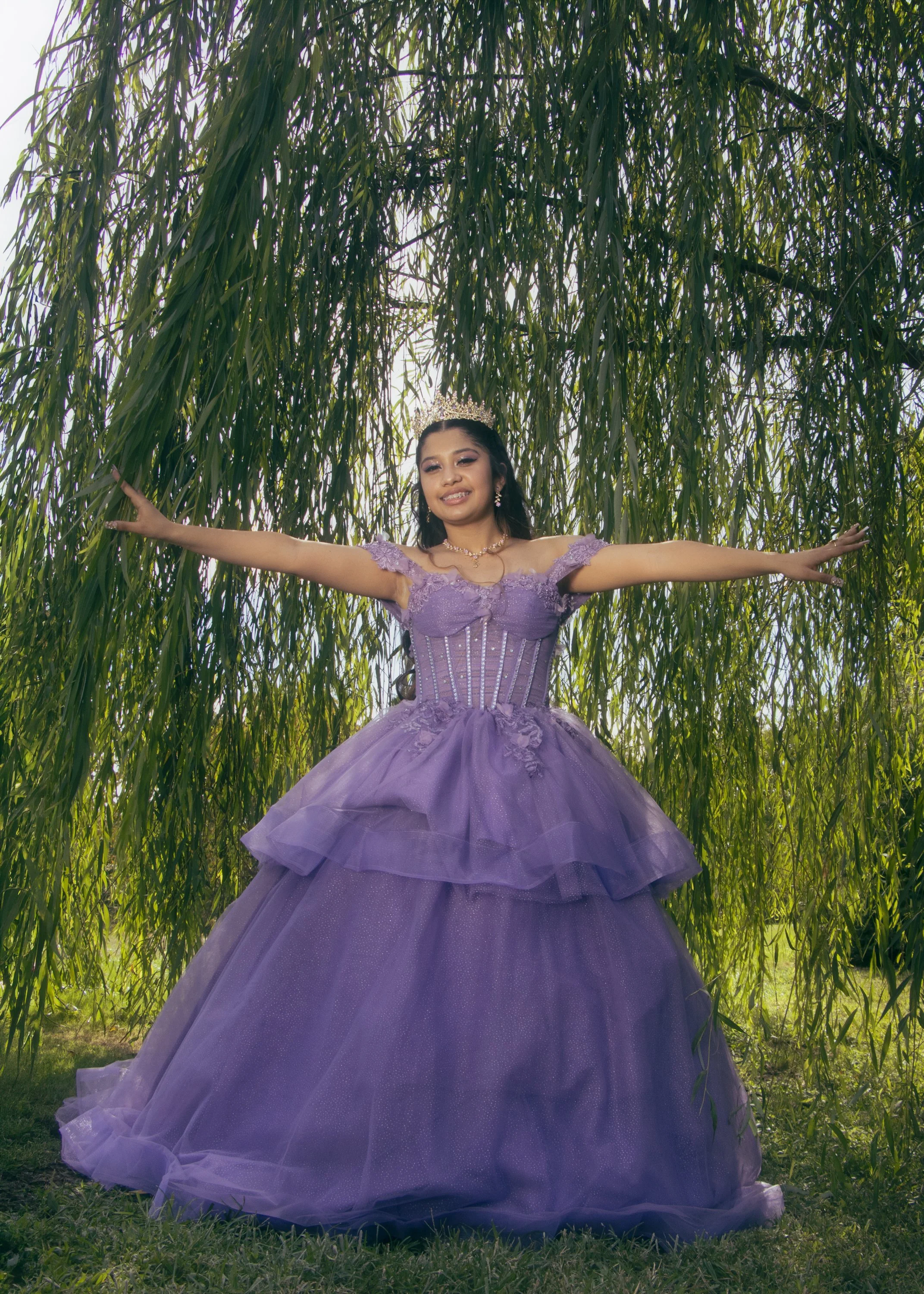 Young woman wearing a lavender princess dress, tiara, and jewelry, standing outdoors under a hanging tree with green foliage, arms extended outward, smiling.
