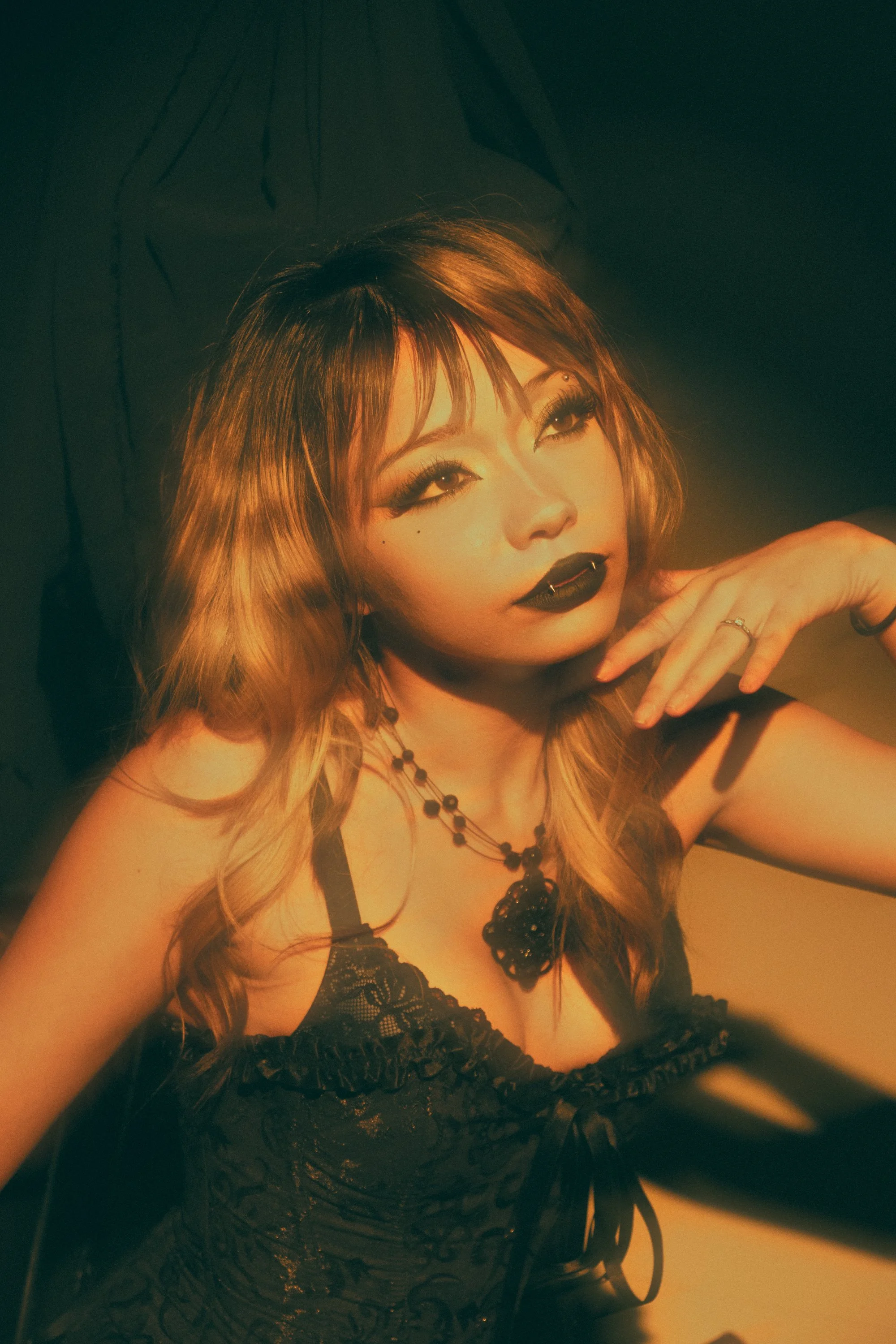 A woman with long, wavy, light brown hair and dark makeup, wearing a black lace top and a necklace with black beads and a dark pendant, posing with her hand near her face against a dark background.