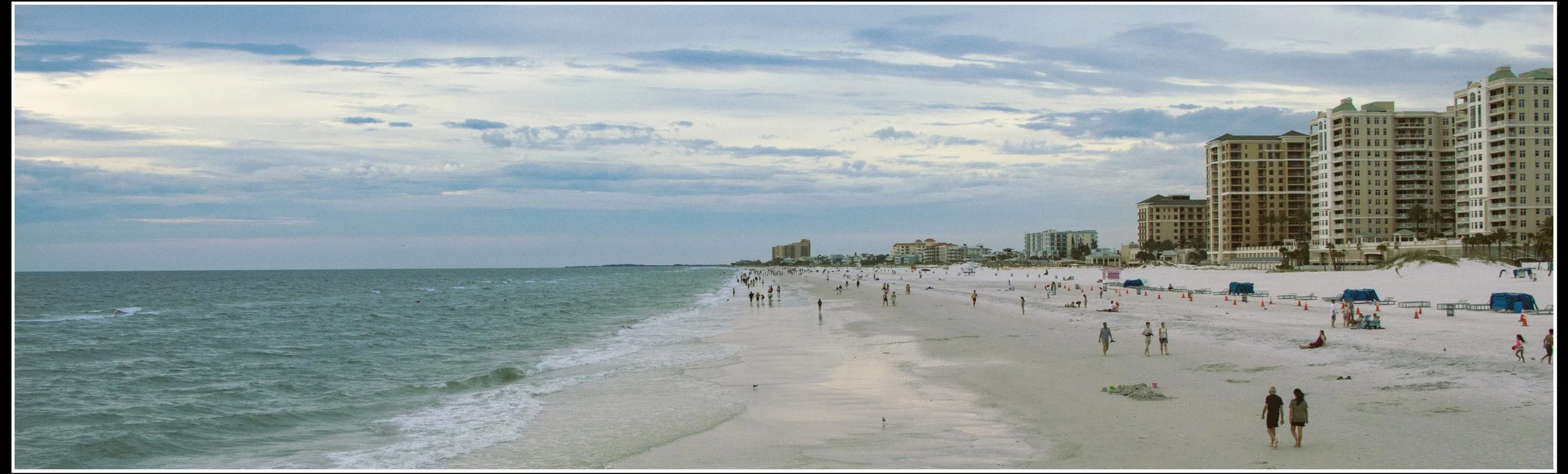 A wide view of a beach with white sand, people walking and relaxing, and high-rise buildings along the shoreline under a cloudy sky.