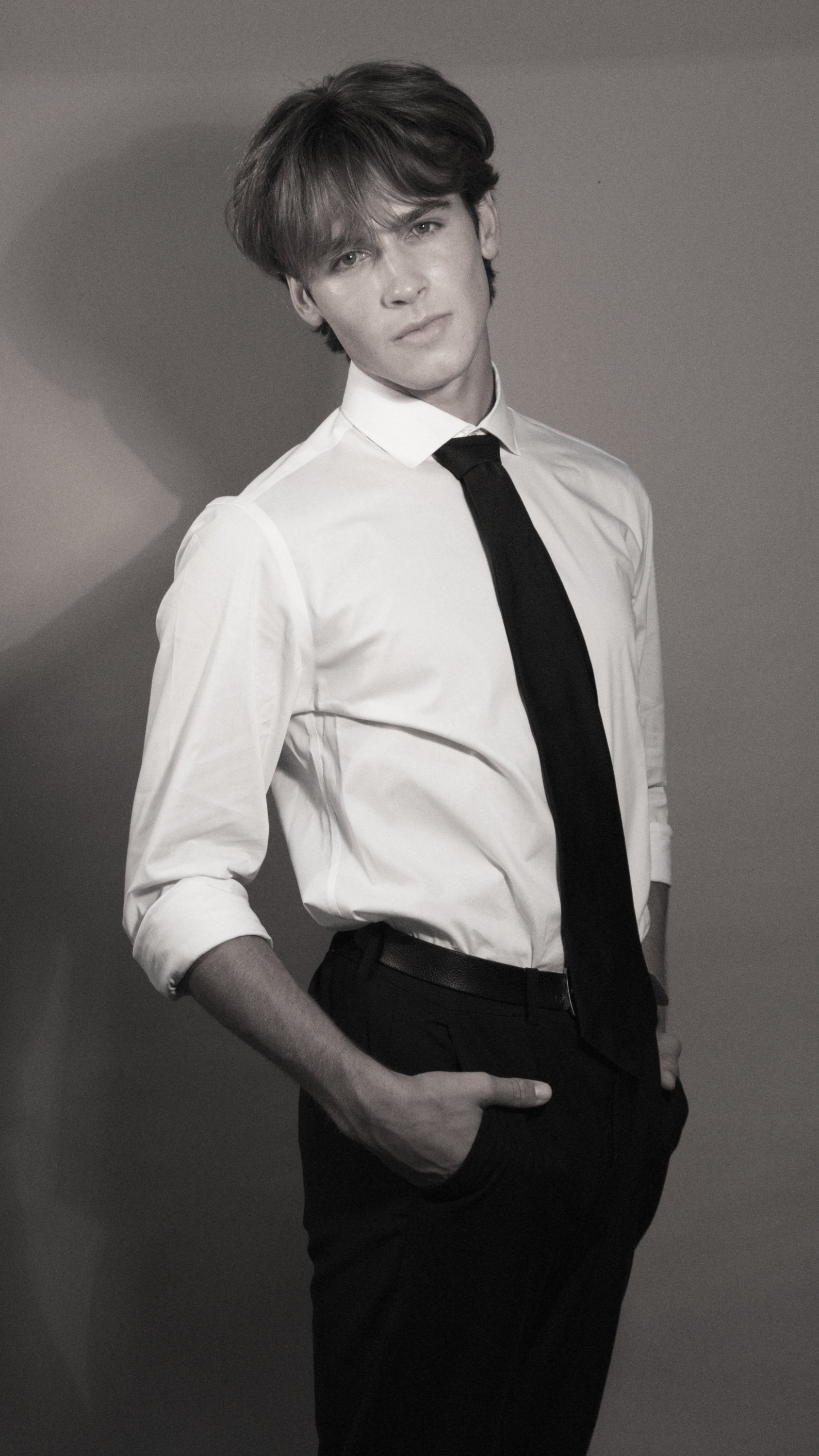 A young man in a white dress shirt, black tie, and black pants stands against a plain background, with his left hand in his pocket. The image is in black and white.