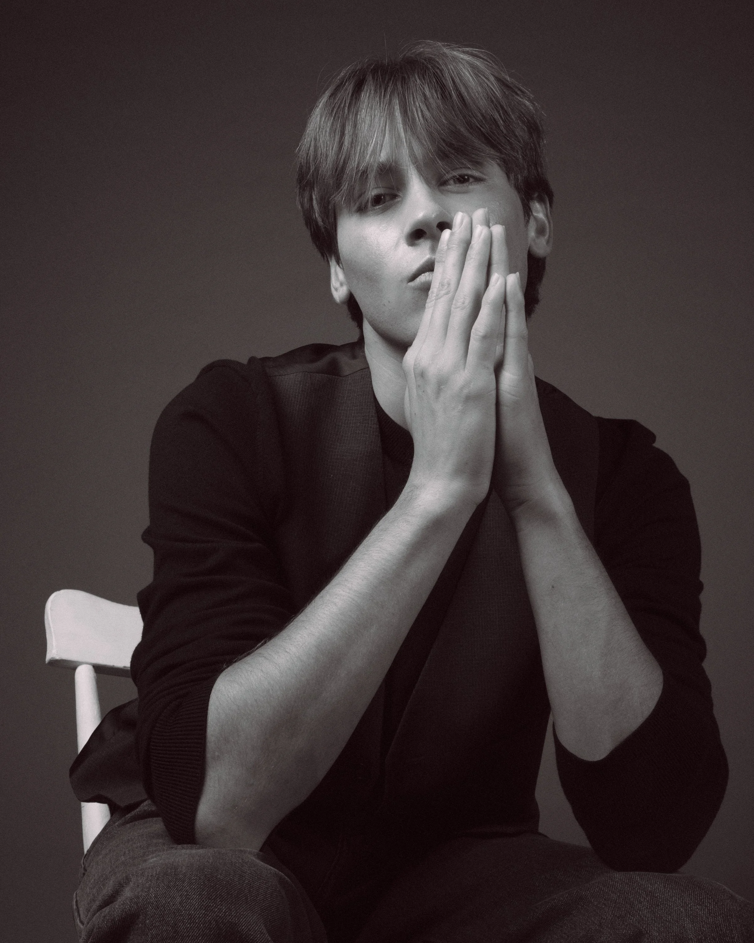 A young man with medium-length hair sitting on a white chair, holding his hands near his face, wearing a black long-sleeve shirt and dark pants, against a dark background.
