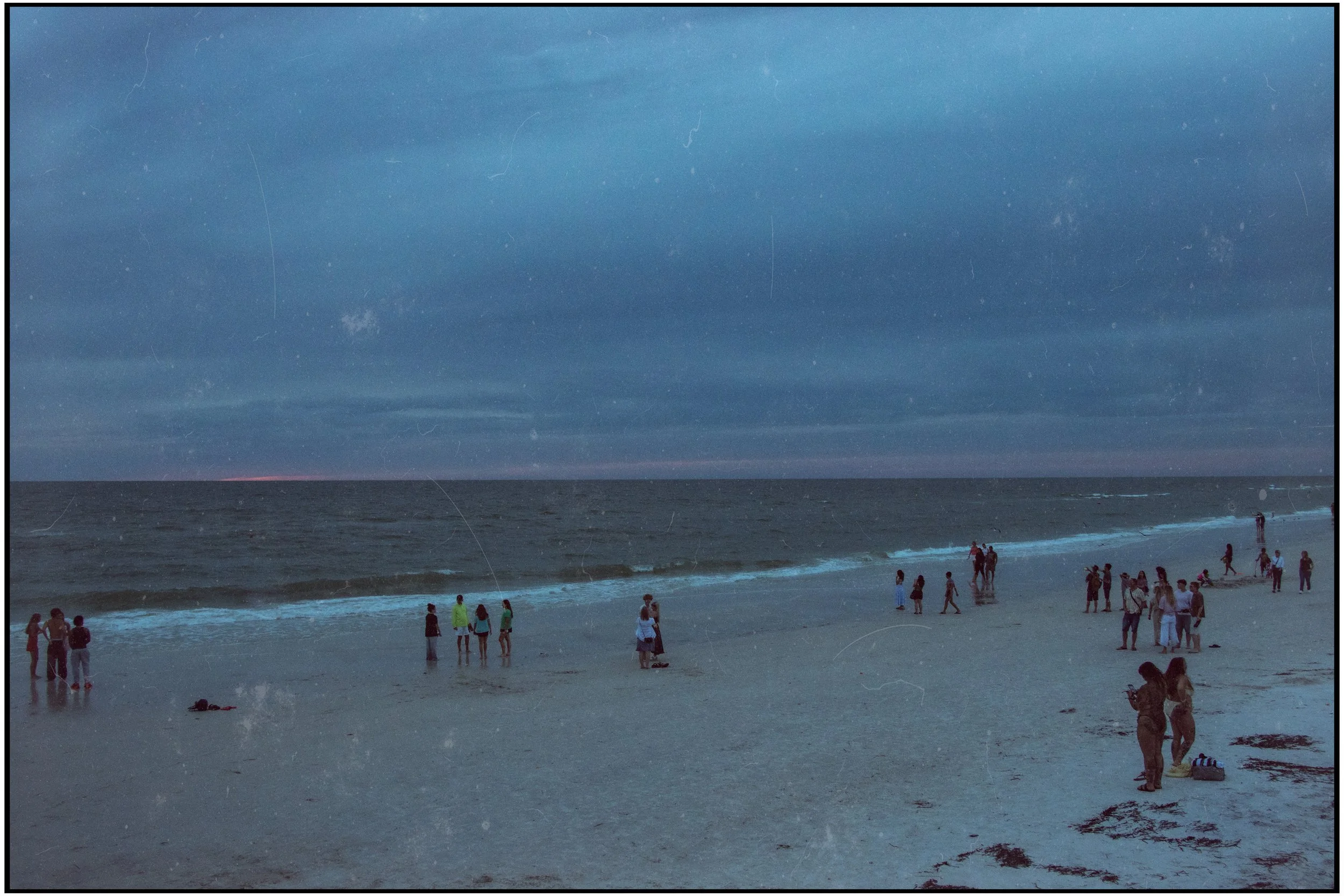 People walking along a beach during sunset with waves crashing and a darkening sky.