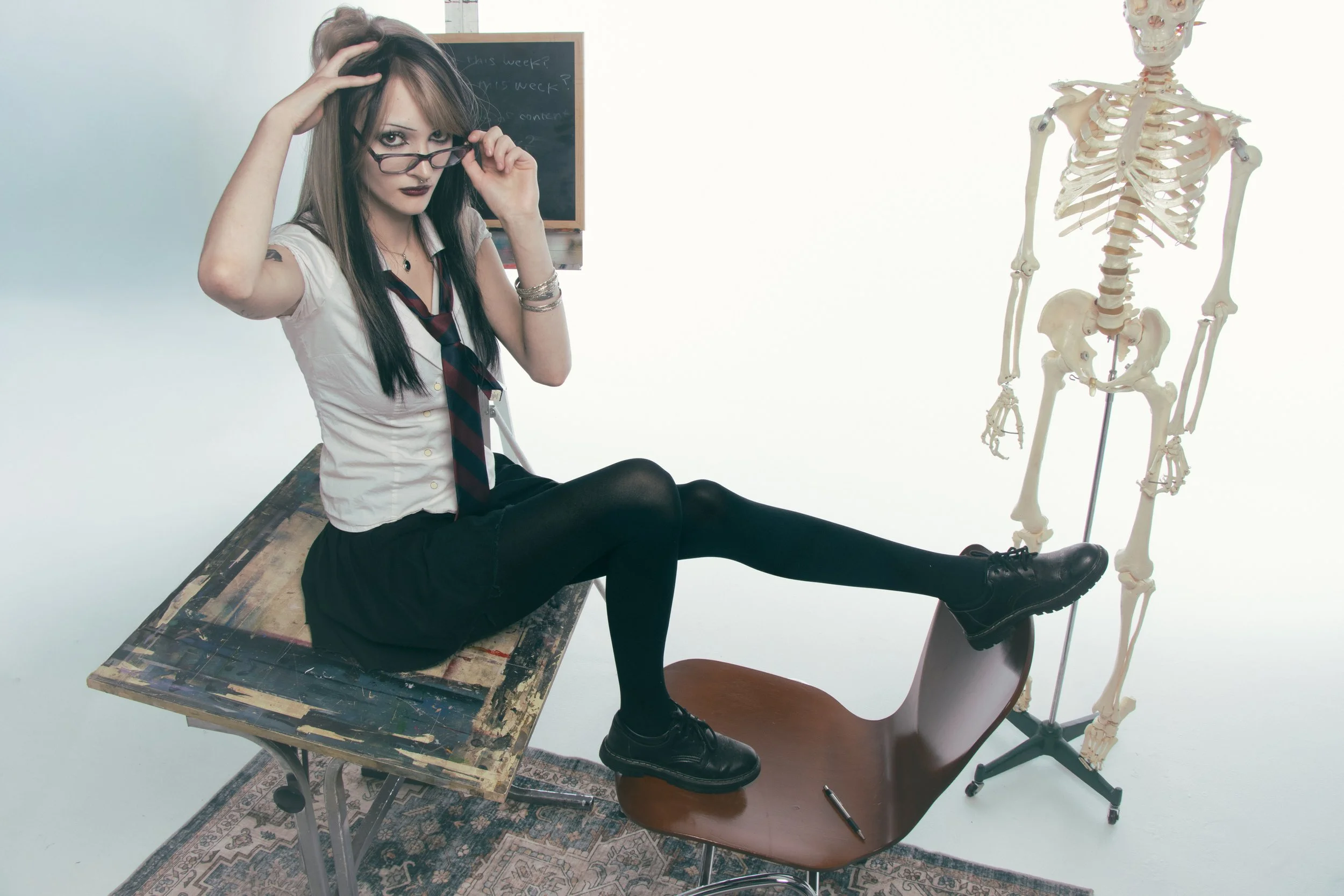 A woman in a school uniform sitting on a table with her foot on a brown chair, looking over her glasses at the camera. A skeleton mannequin is beside her in a studio setting, with a chalkboard in the background.