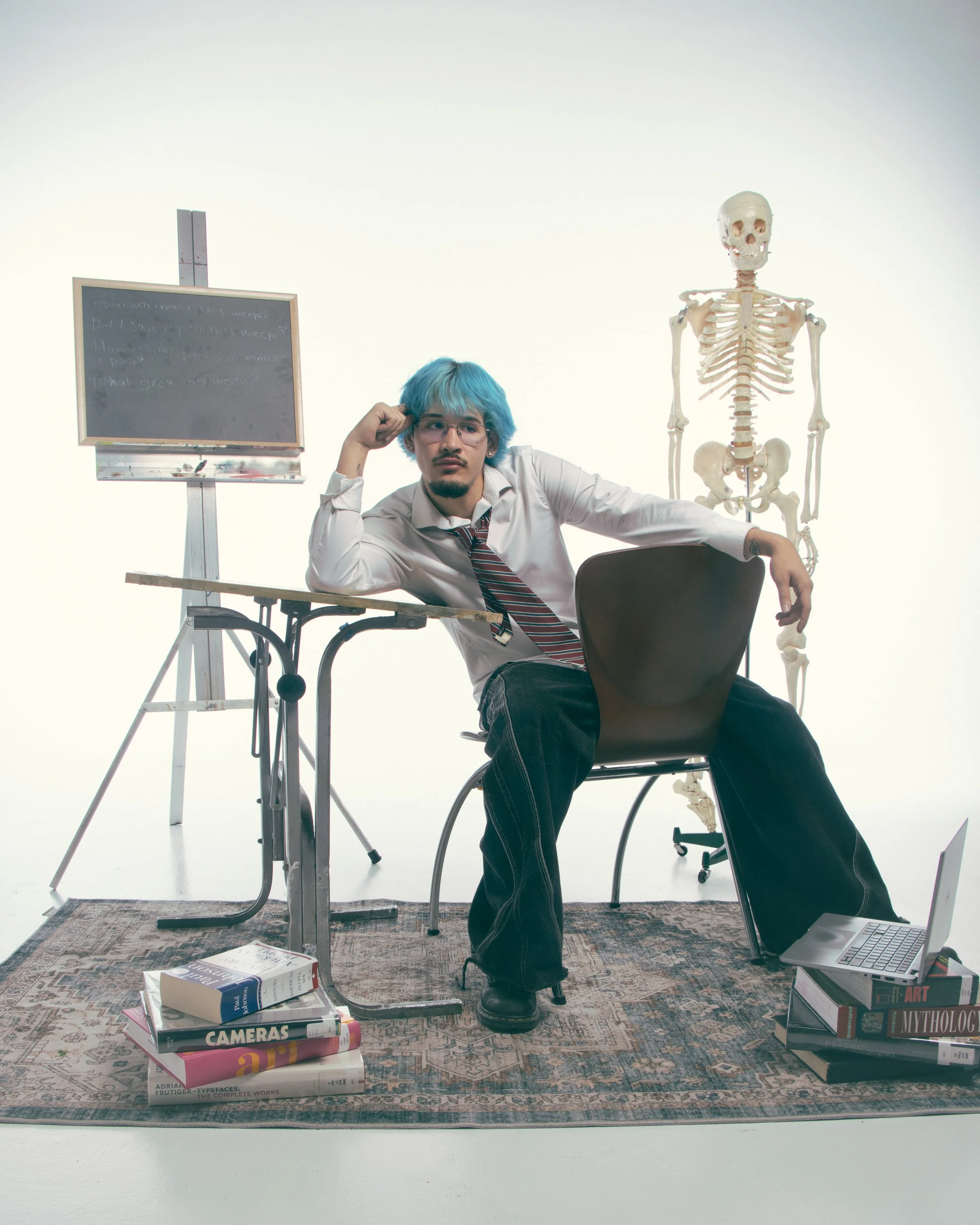 A person with blue hair and glasses sitting at a desk, with a skeleton model to their right, a stack of books on the floor, and more books stacked on the chair, in a room with light background and a black chalkboard on a stand.
