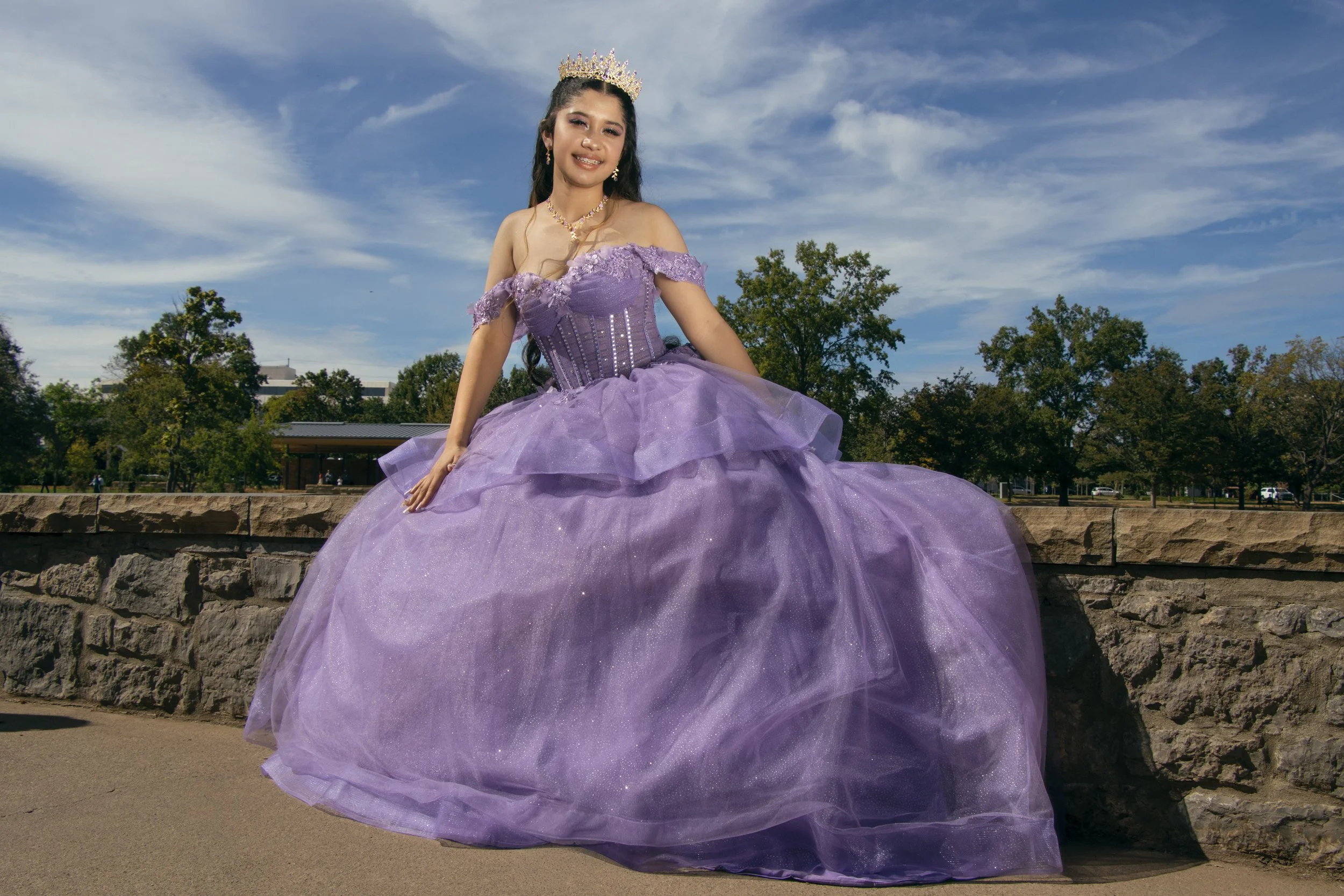 A young woman dressed in a purple ball gown with a crown on her head, standing outdoors against a backdrop of trees and a partly cloudy sky.