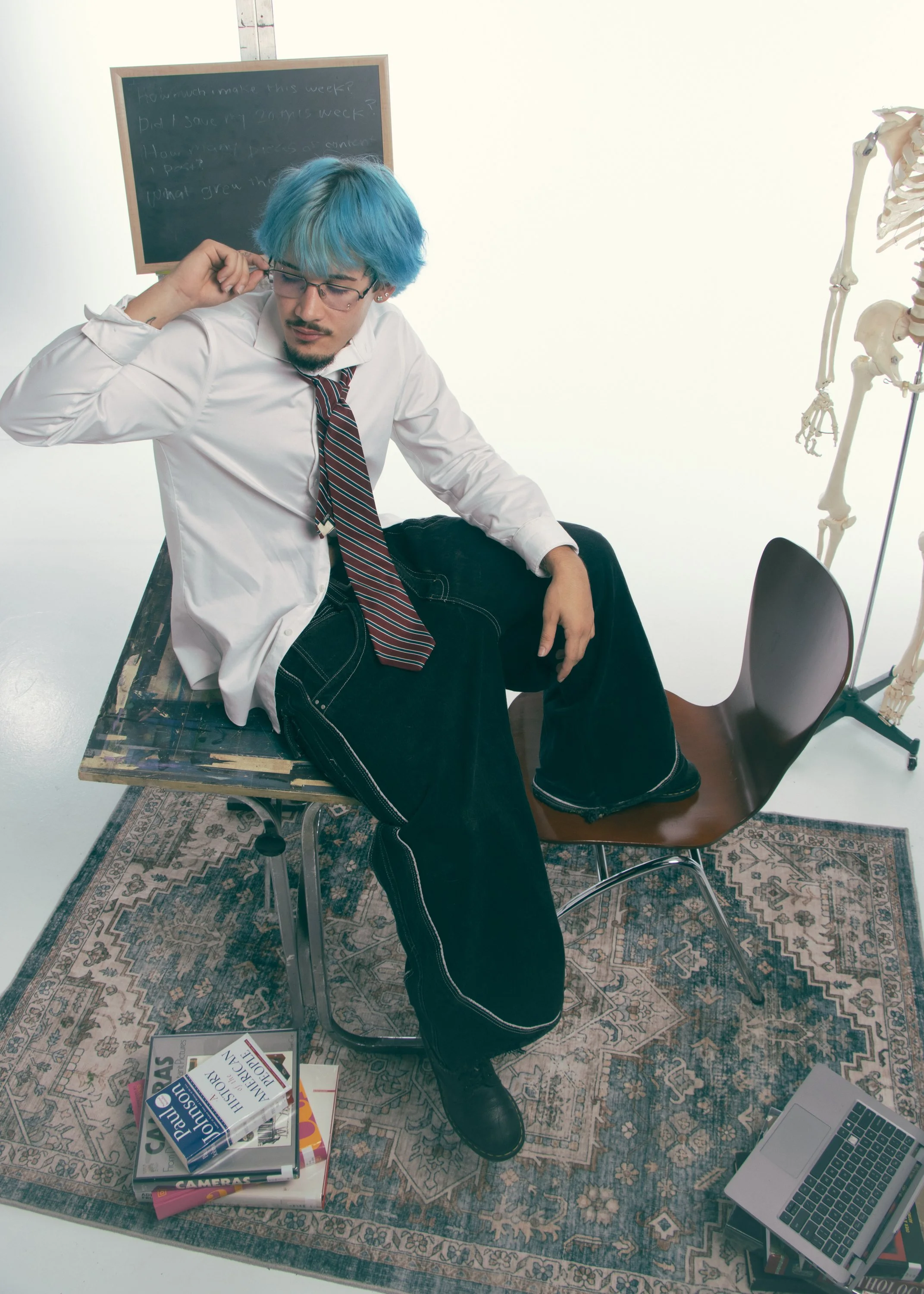 A man with blue hair and glasses sitting on a wooden chair, leaning back with one arm resting on a table. The table has several books and a laptop on it. There is a skeleton to the right and a chalkboard behind him with writing on it. The man is dres