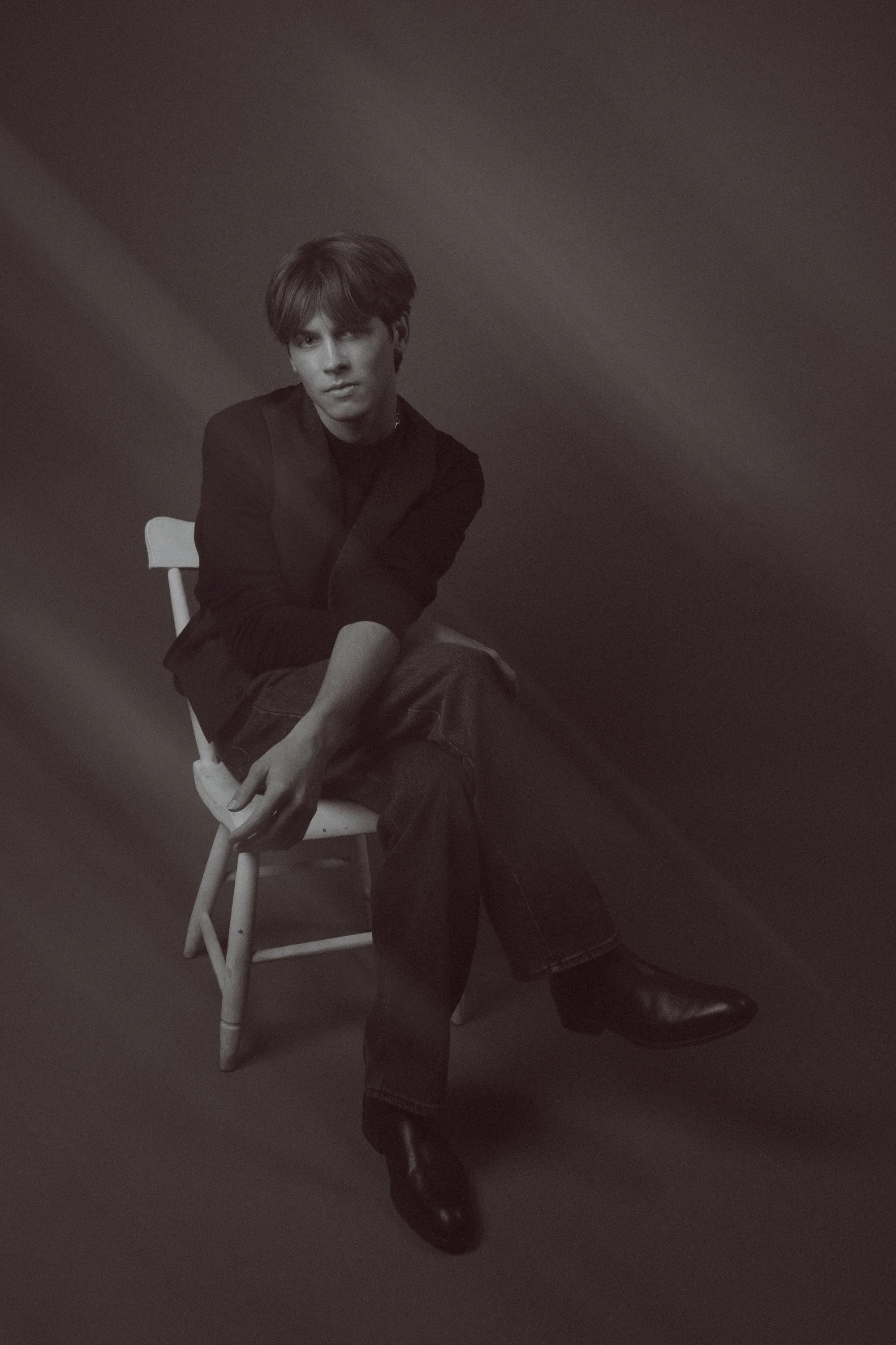 A young man with dark hair sitting on a white chair against a plain dark wall, dressed in dark clothing, with one leg crossed over the other and his hands resting on his knee.