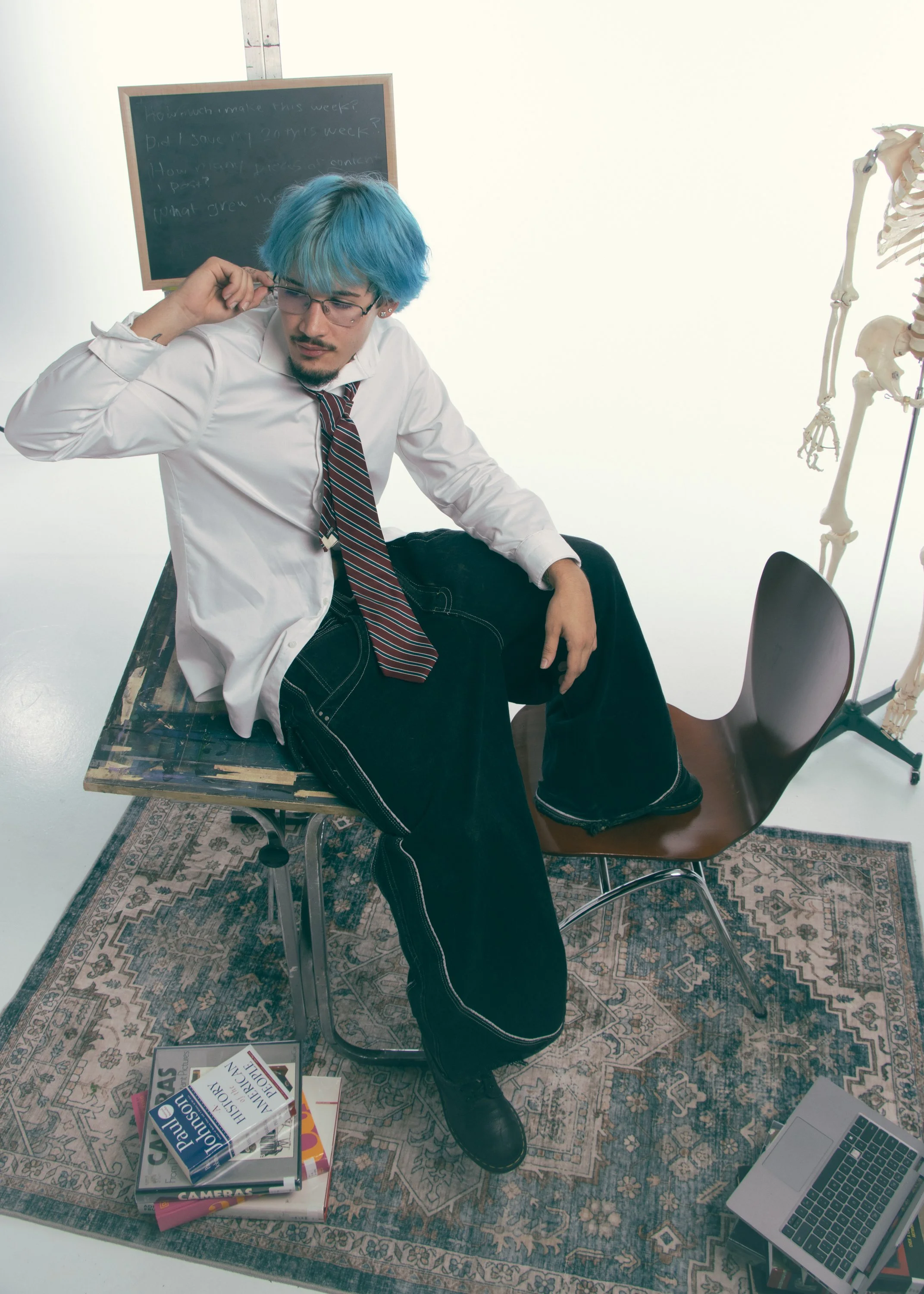 A man with blue hair, glasses, and a striped tie sits on a chair with one leg crossed over the other in a room with a patterned rug, a skeleton model, and a stack of books on photography and cameras. A textbook on digital photography is near the book
