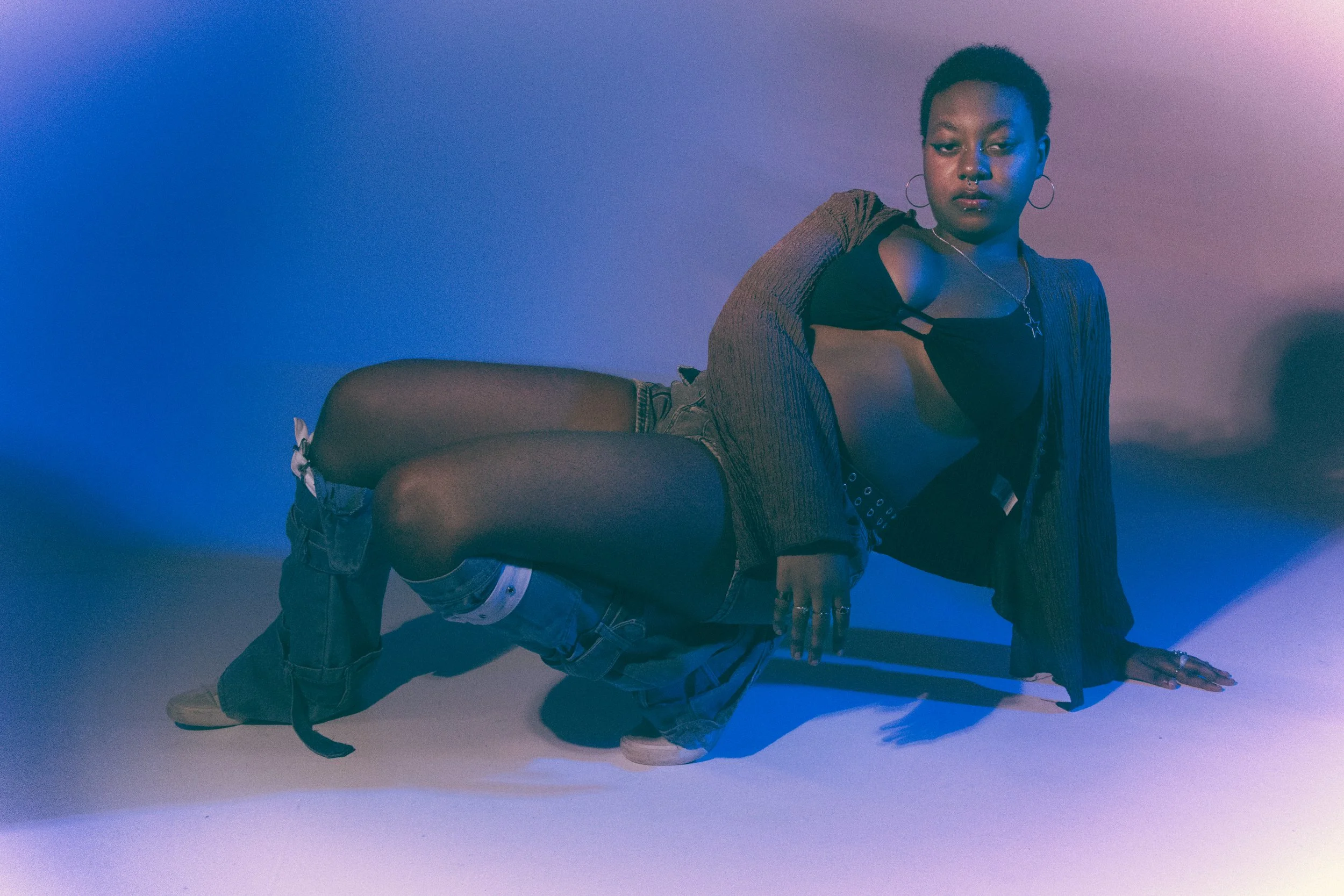 A woman with short hair and hoop earrings poses on the floor in front of a blue and purple gradient background. She wears a black top, a brown cardigan, shorts, and sneakers with white socks, and has a necklace with a star pendant.
