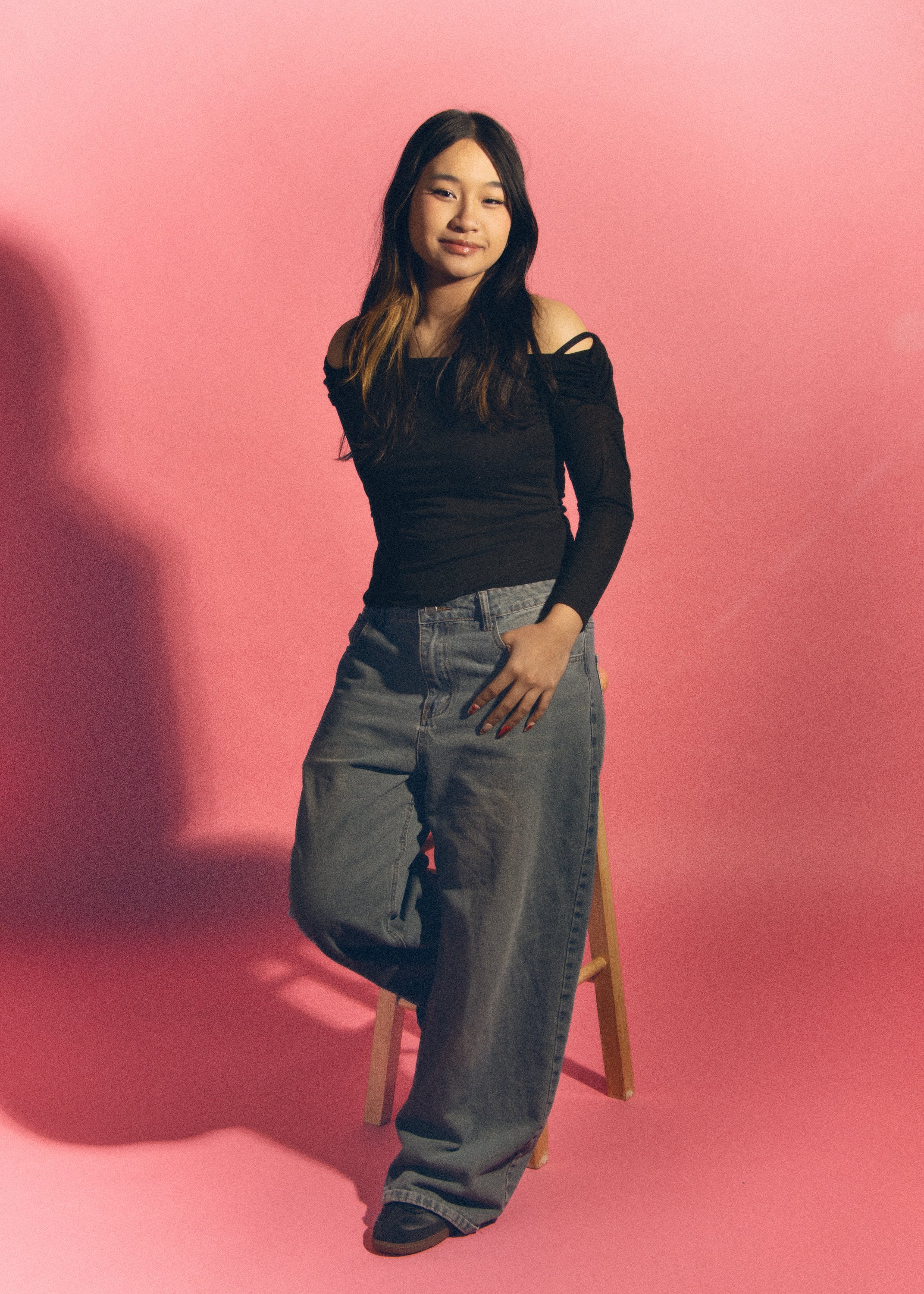 A young woman with long dark hair styled with light brown highlights, wearing an off-shoulder black top and baggy gray jeans, standing in front of a pink background with a wooden chair partially visible behind her.