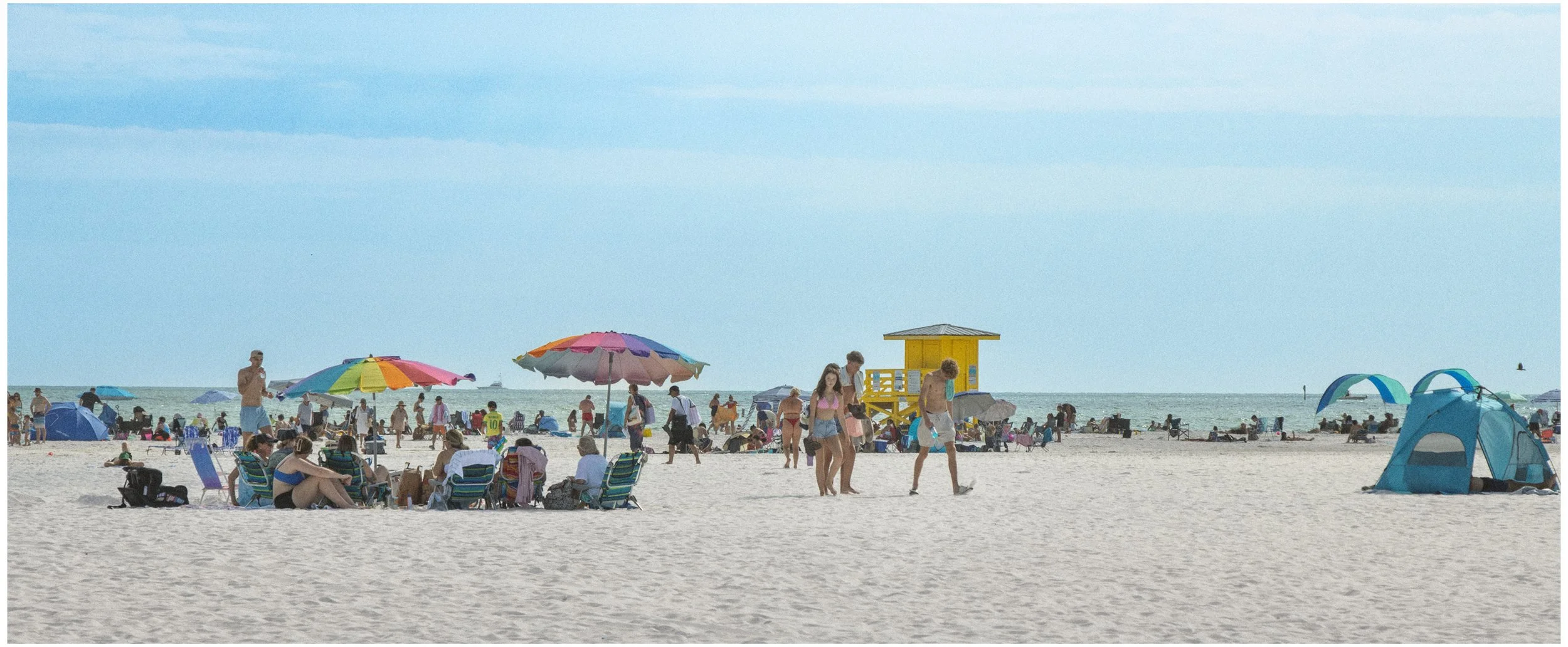 A crowded beach scene with umbrellas, tents, people relaxing, and walking along the sand. Lifeguard tower and ocean in the background.