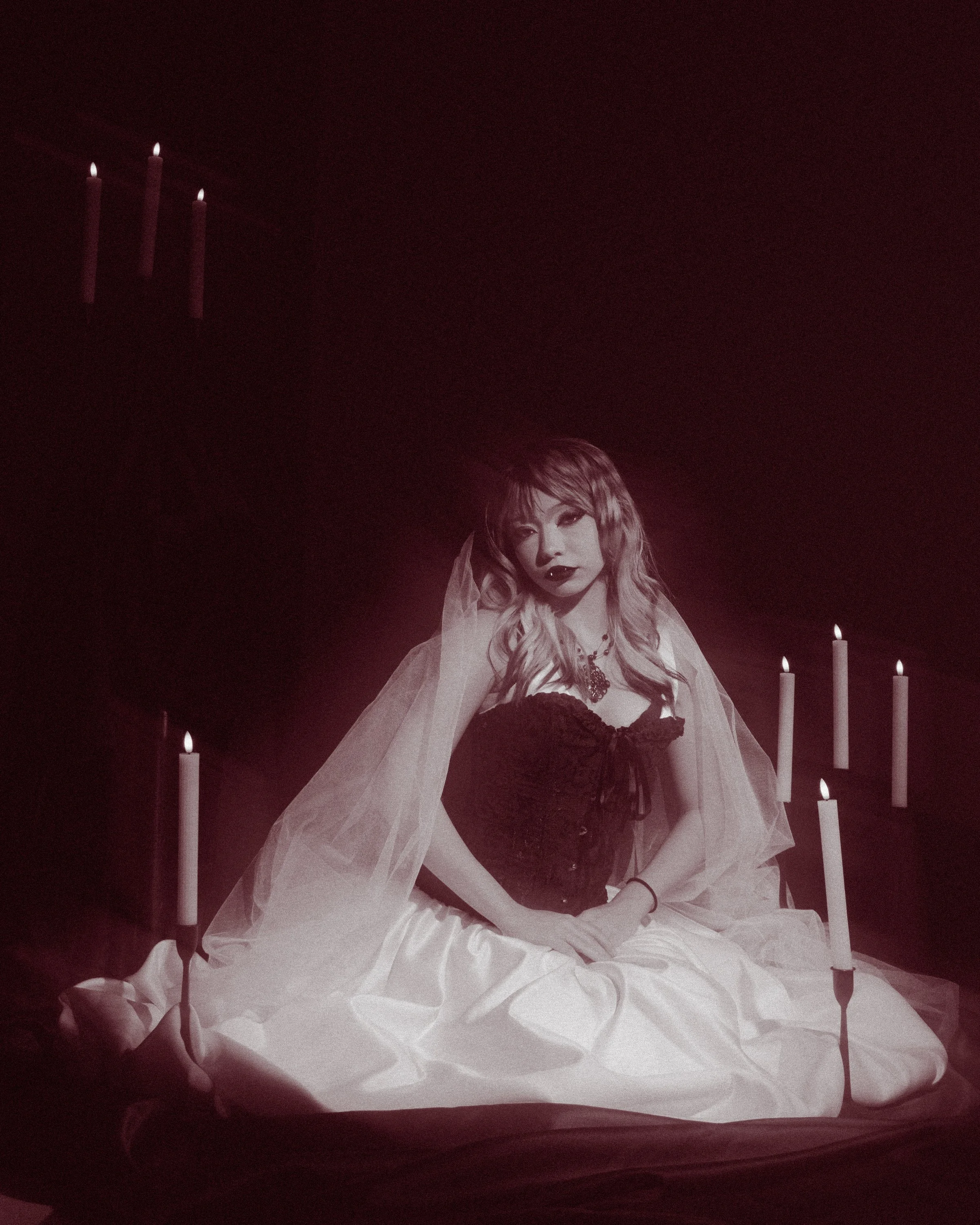 Black-and-white photo of a woman with wavy hair, wearing makeup, a dark corset, a veil, and jewelry, surrounded by lit candles.