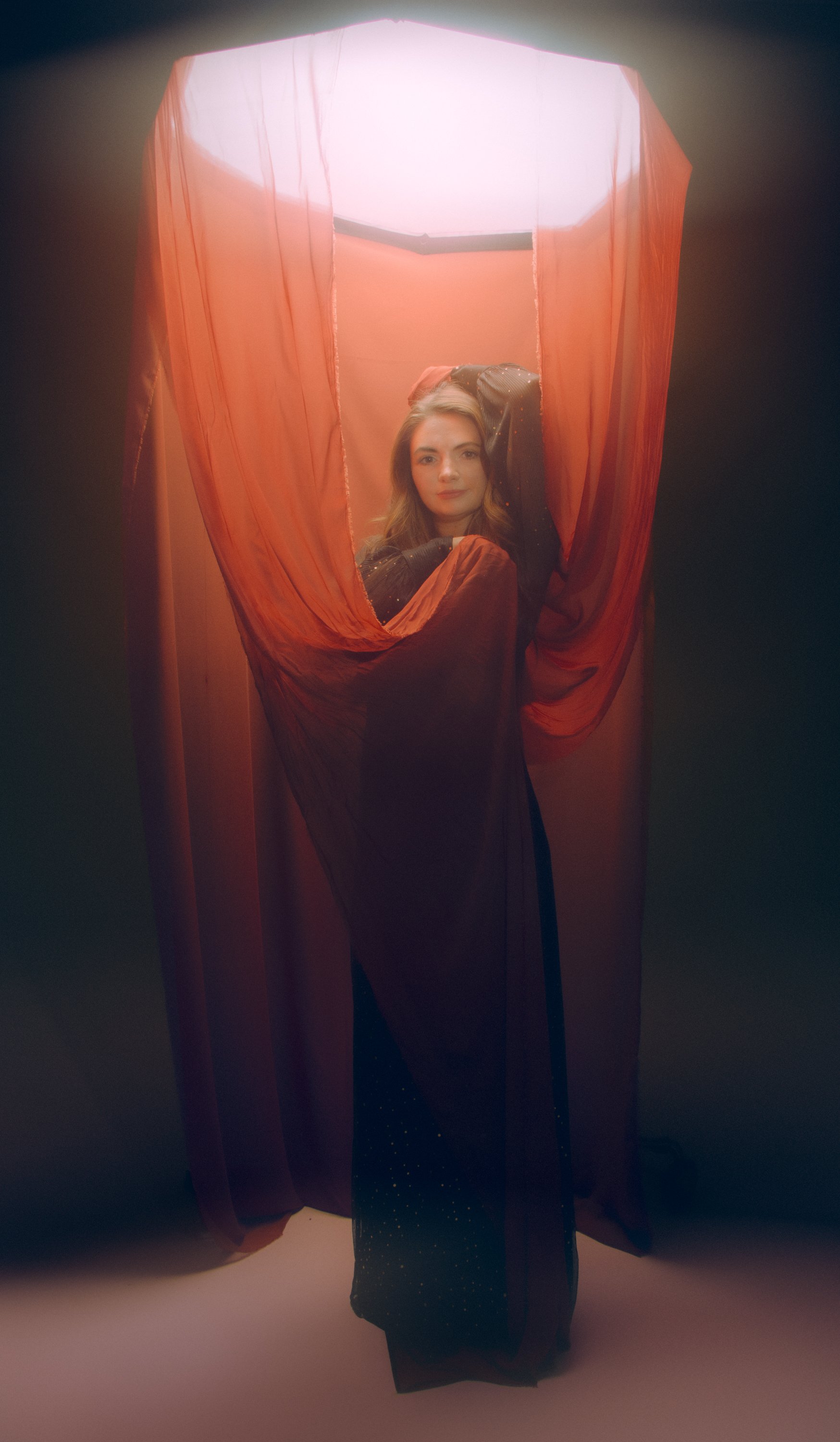 A young woman with long hair standing behind a sheer red curtain, looking directly at the camera with her hand resting behind her head, illuminated by soft light from above.