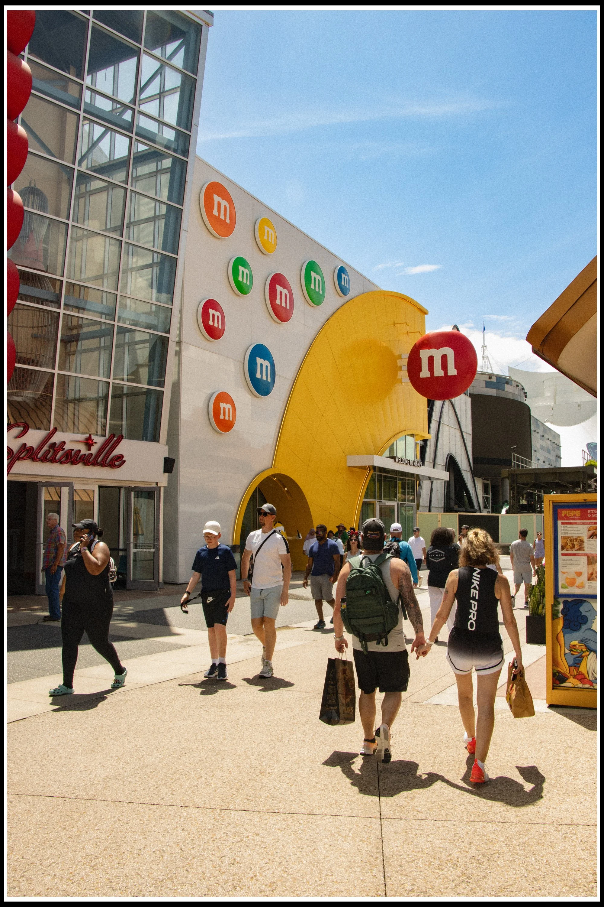 People walking outside the M&M's World store on a sunny day. The store has colorful M&Ms logos on the wall and large red M&M character balloons.