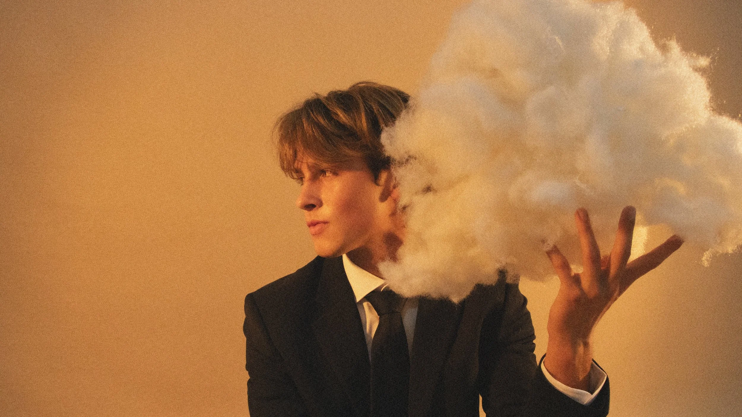 A young man in a black tuxedo and bowtie is holding a cloud of vapor or smoke near his face, making a peace sign with his fingers against a warm beige background.