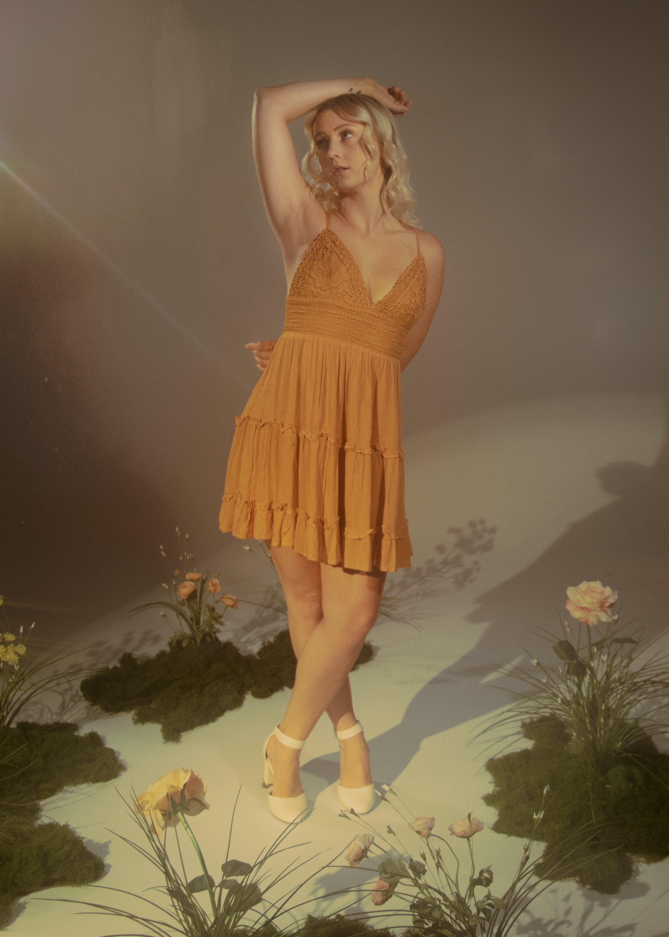 A woman with blonde curly hair posing in a yellow lace dress and white heels in a studio with flowers and greenery on the ground.