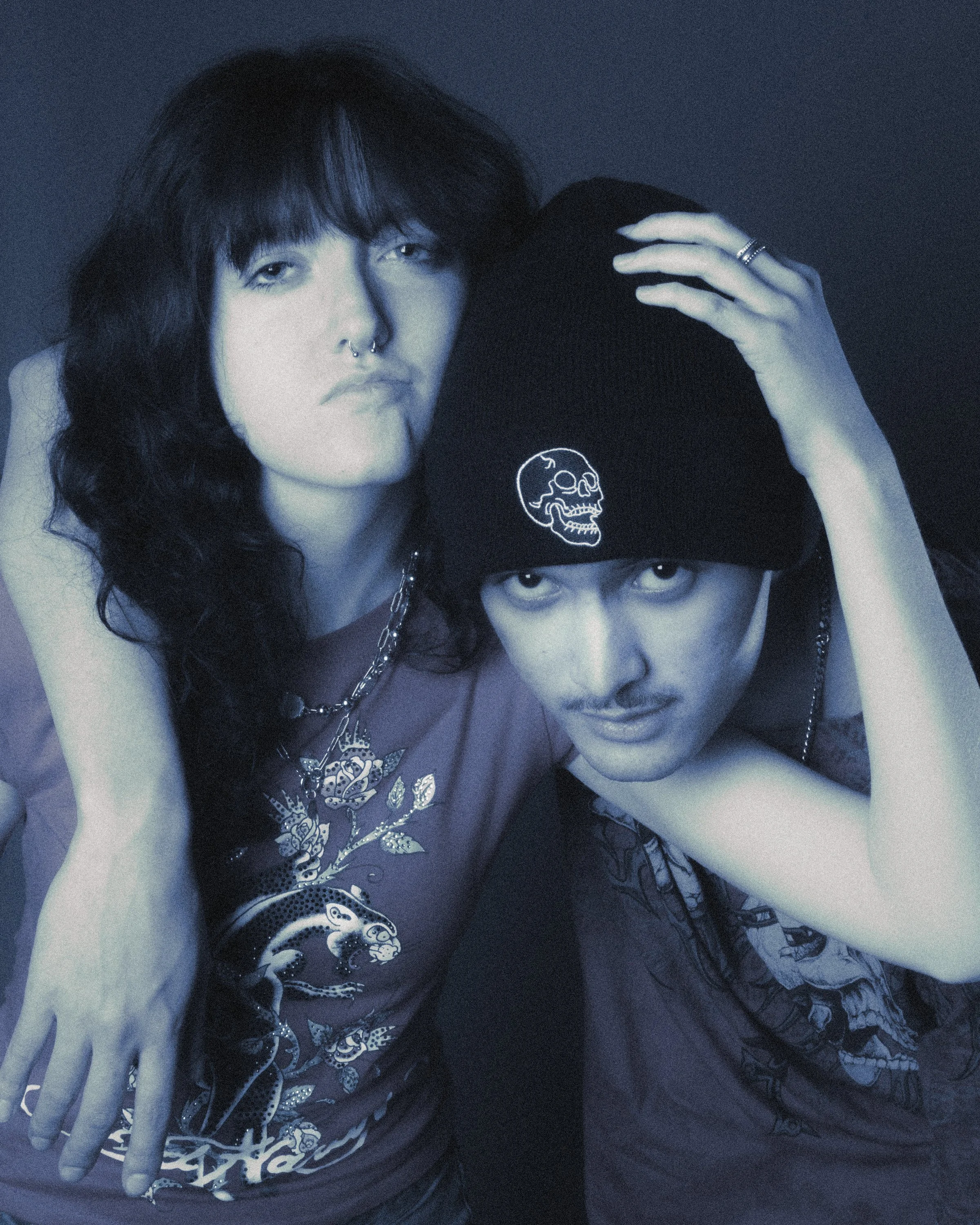 Black and white photo of two people. One person has long, dark hair, multiple rings on fingers, and a septum piercing. The other person wears a black cap with a white skull and snake design and has facial tattoos, with their eyes visible. The person 