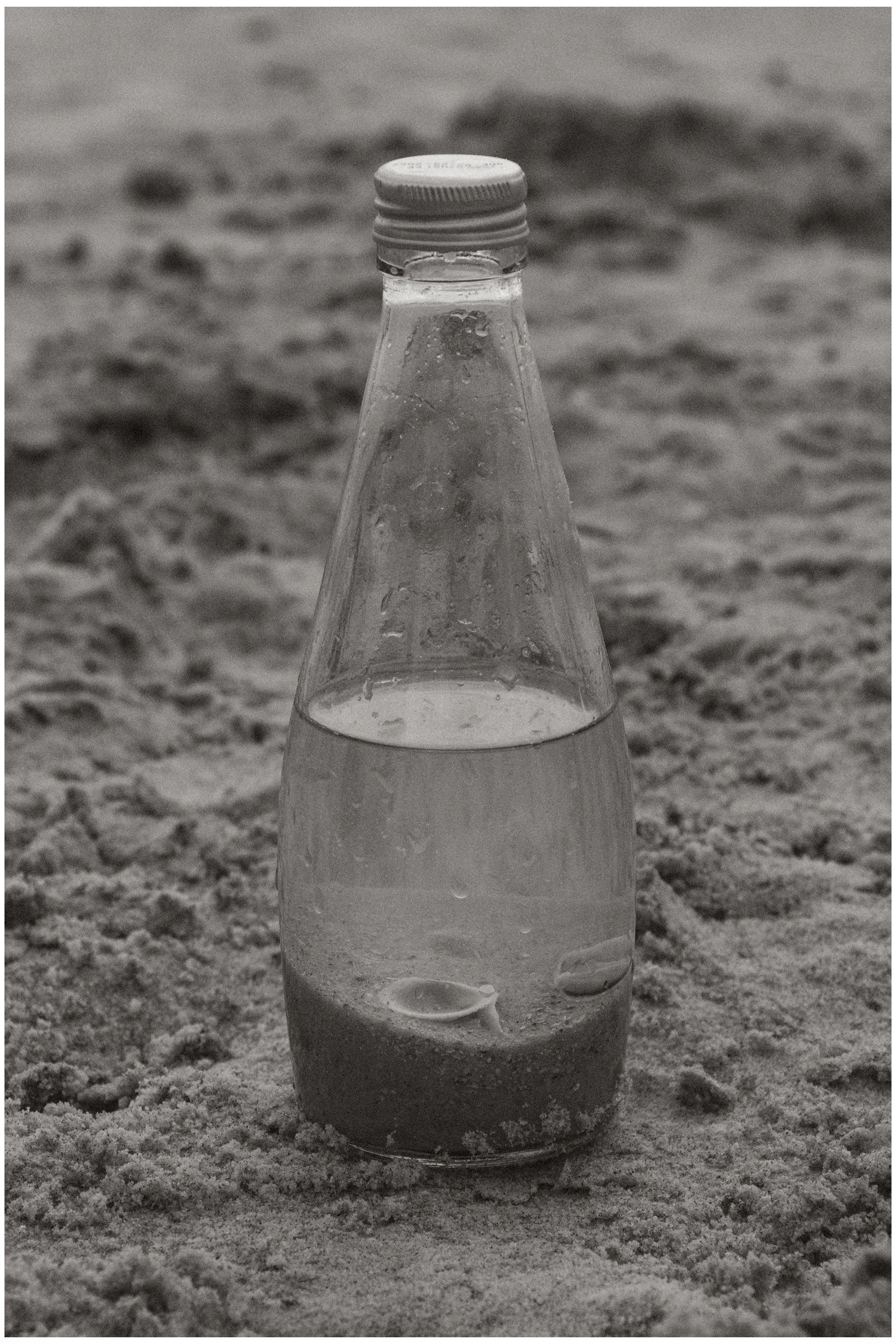 A clear glass bottle with a white cap, filled with a transparent liquid, placed on sandy ground in an outdoor setting.