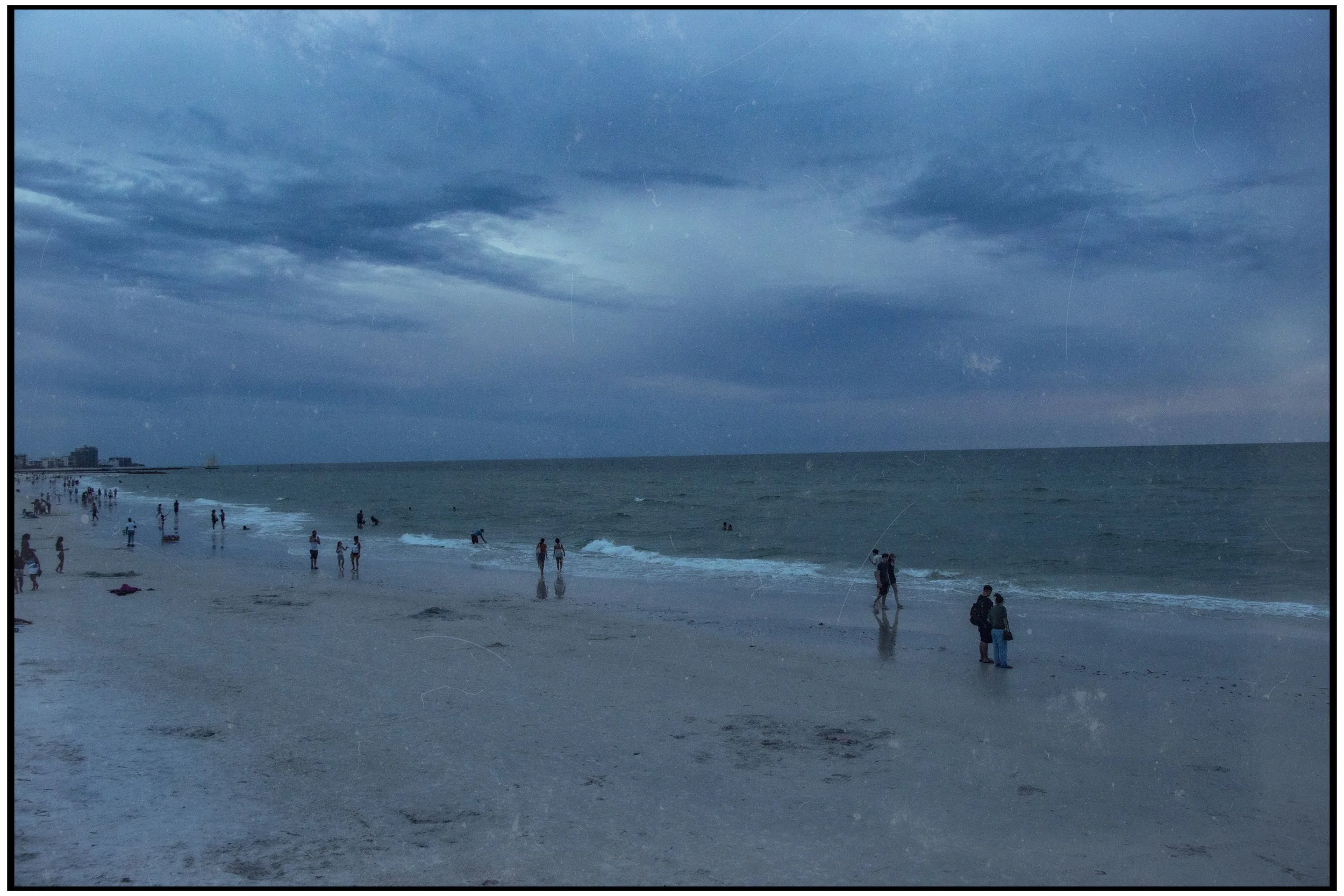 Overcast sky with dark clouds over a busy beach with people walking and swimming in the water, some buildings visible in the distance.