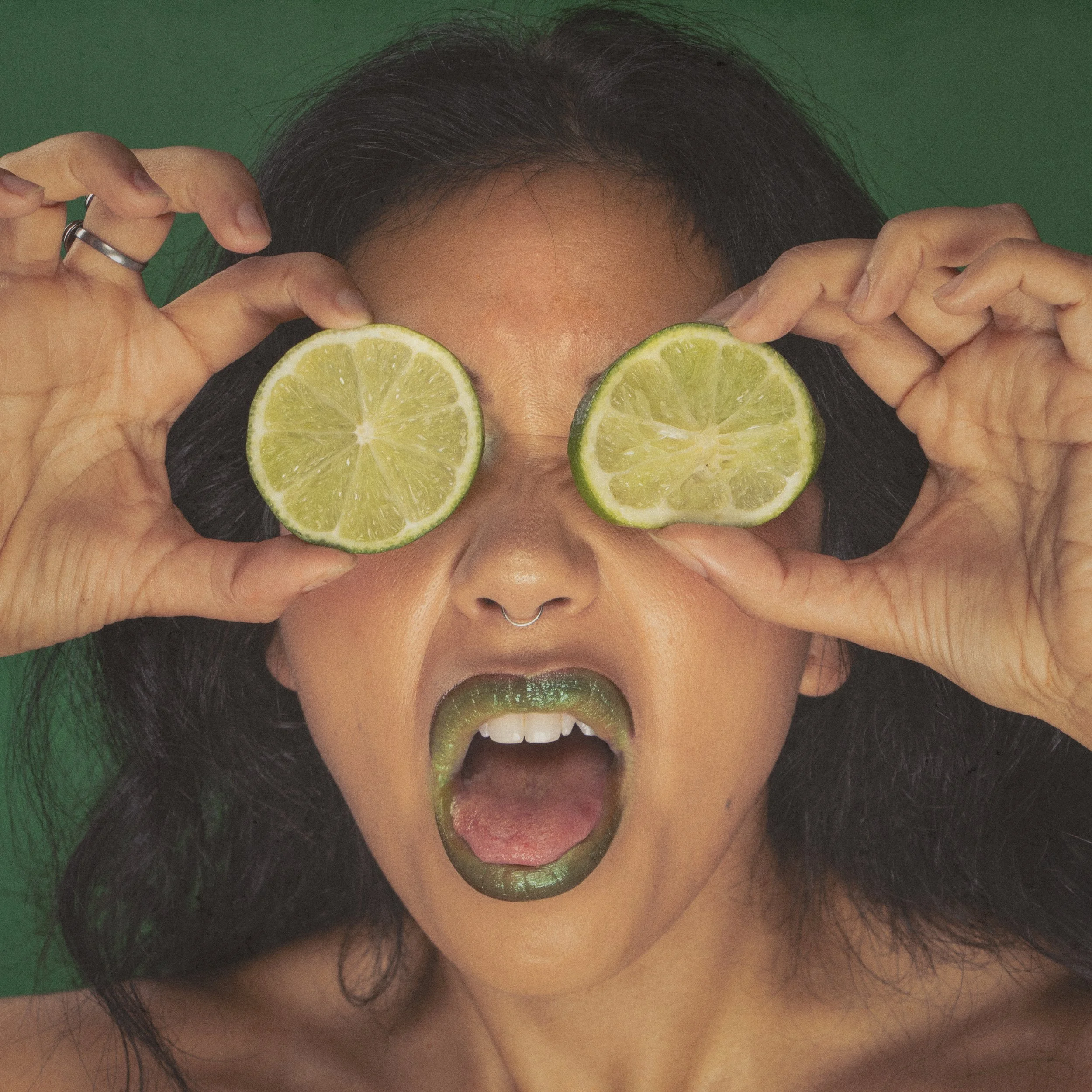 A person with black hair and a nose ring holding lime slices in front of their eyes, showing only their forehead, nose, mouth, and chin against a green background.