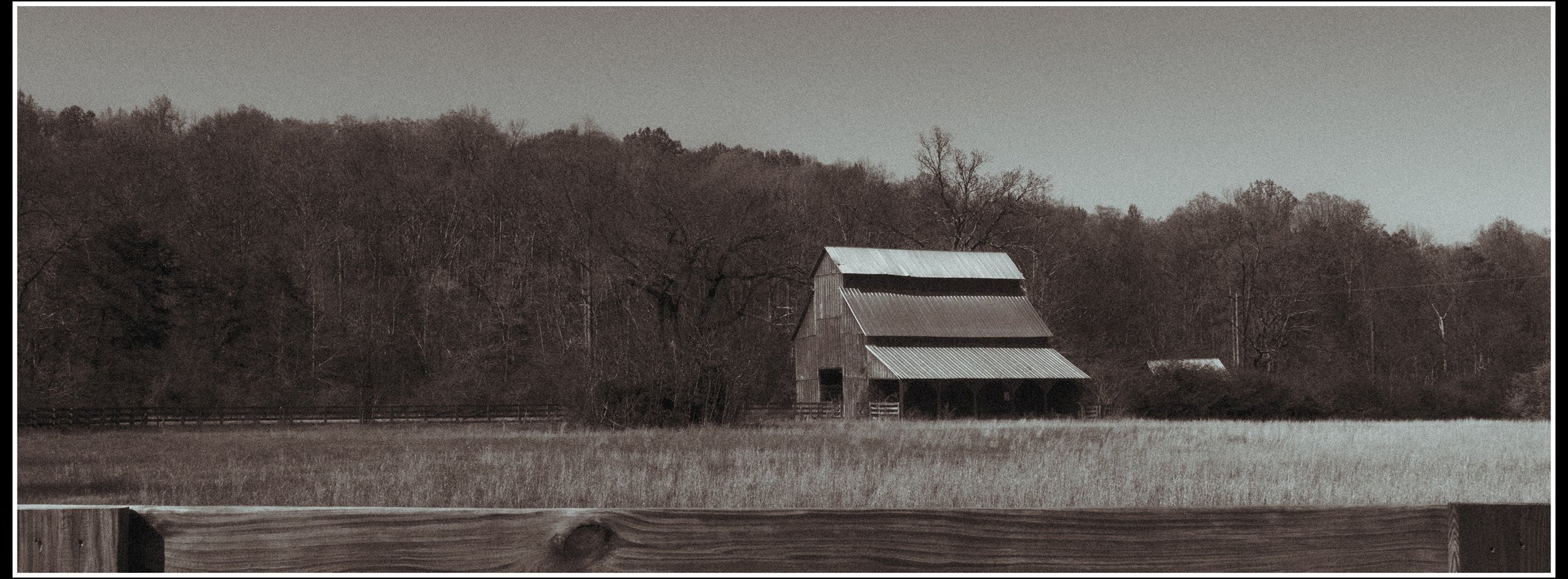 A rural landscape with a weathered wooden barn and trees in the background, viewed across a grassy field.