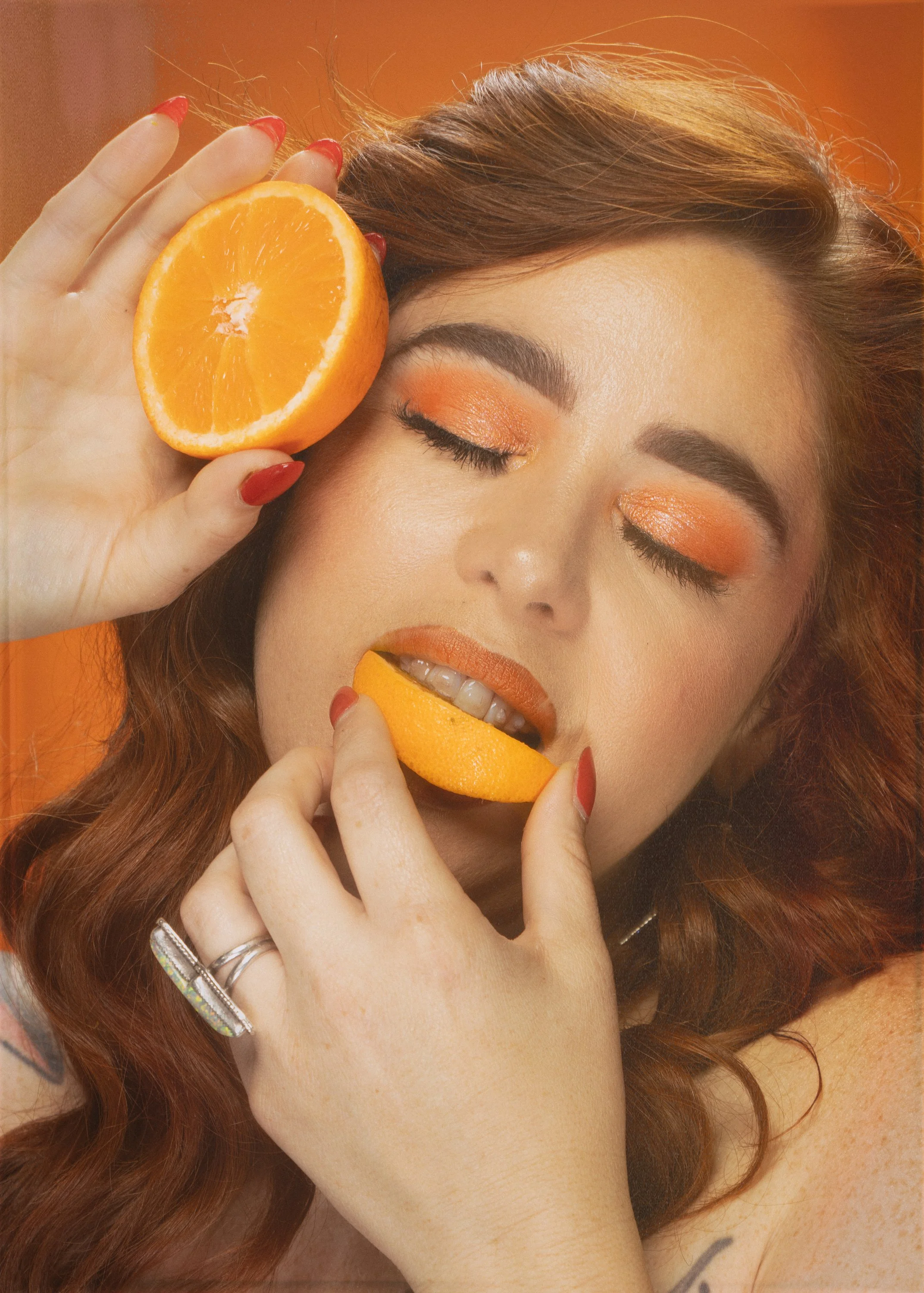 A woman with red wavy hair and makeup holding a halved orange near her face and a slice in her mouth, with a warm orange background.