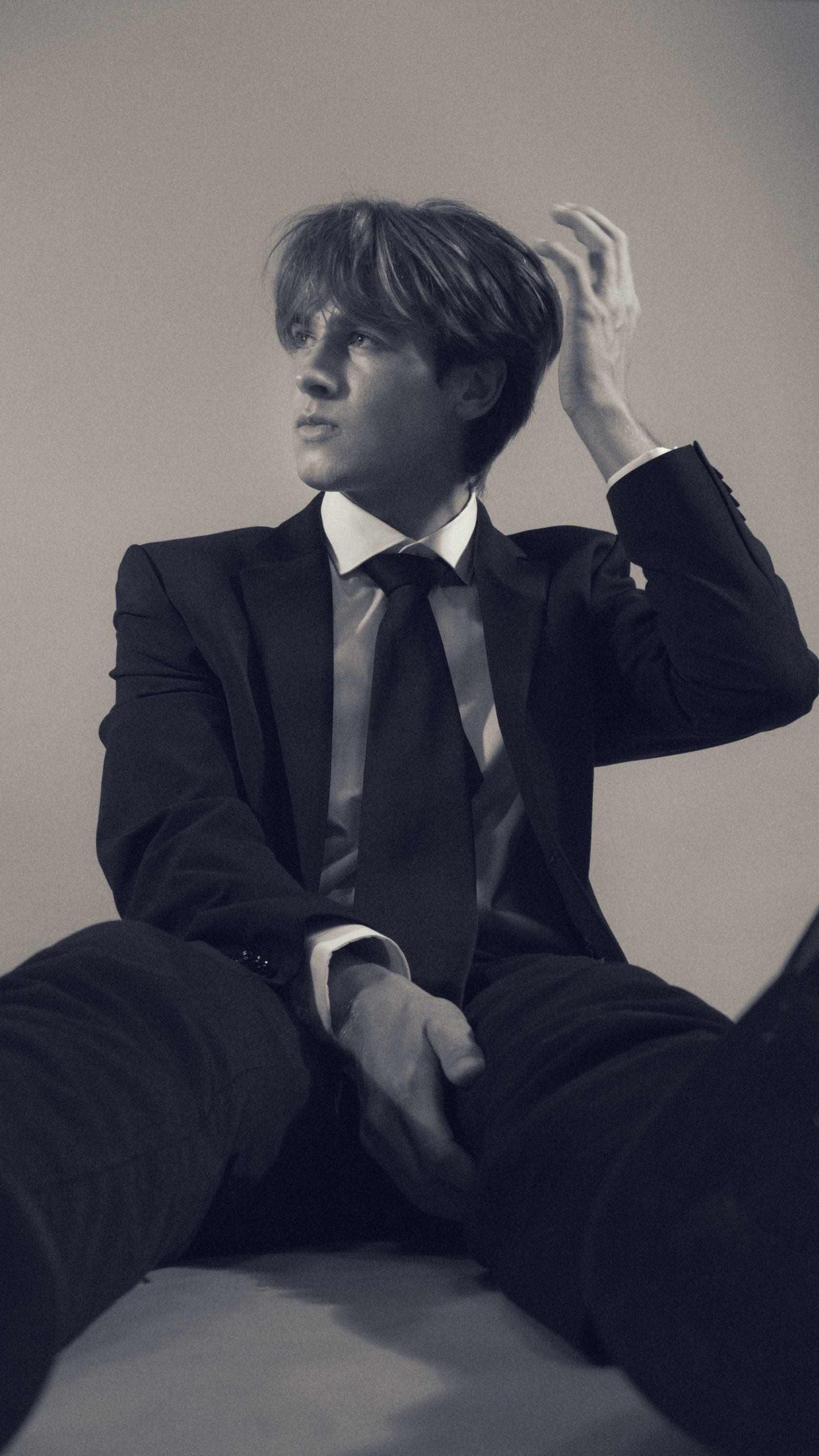 Black-and-white photo of a young man in a suit sitting on the floor, with one hand on his head, looking away from the camera.