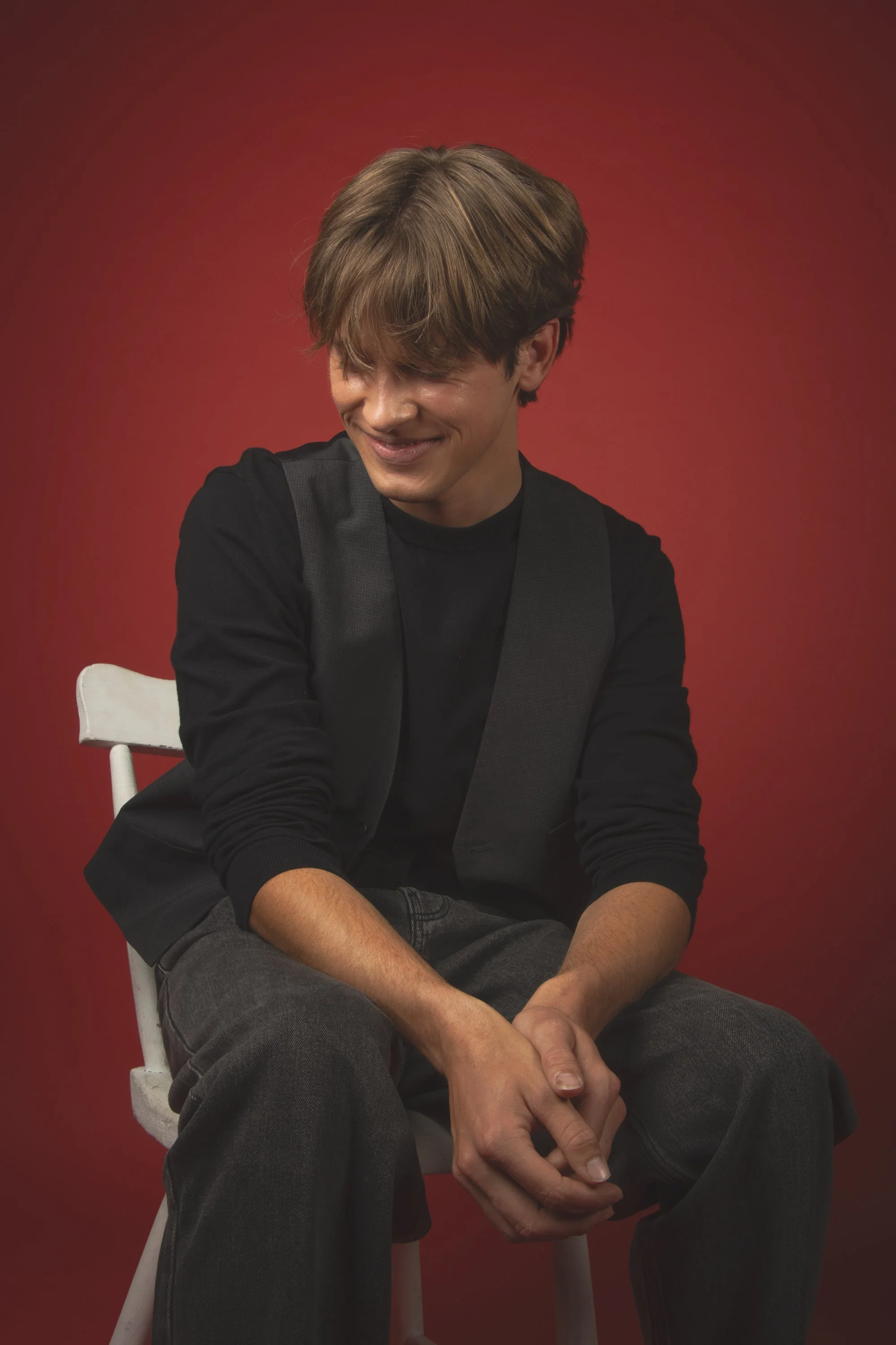 A young man with brown hair wearing a black shirt and dark pants is sitting on a white chair against a red background, smiling with his eyes closed and hands clasped.