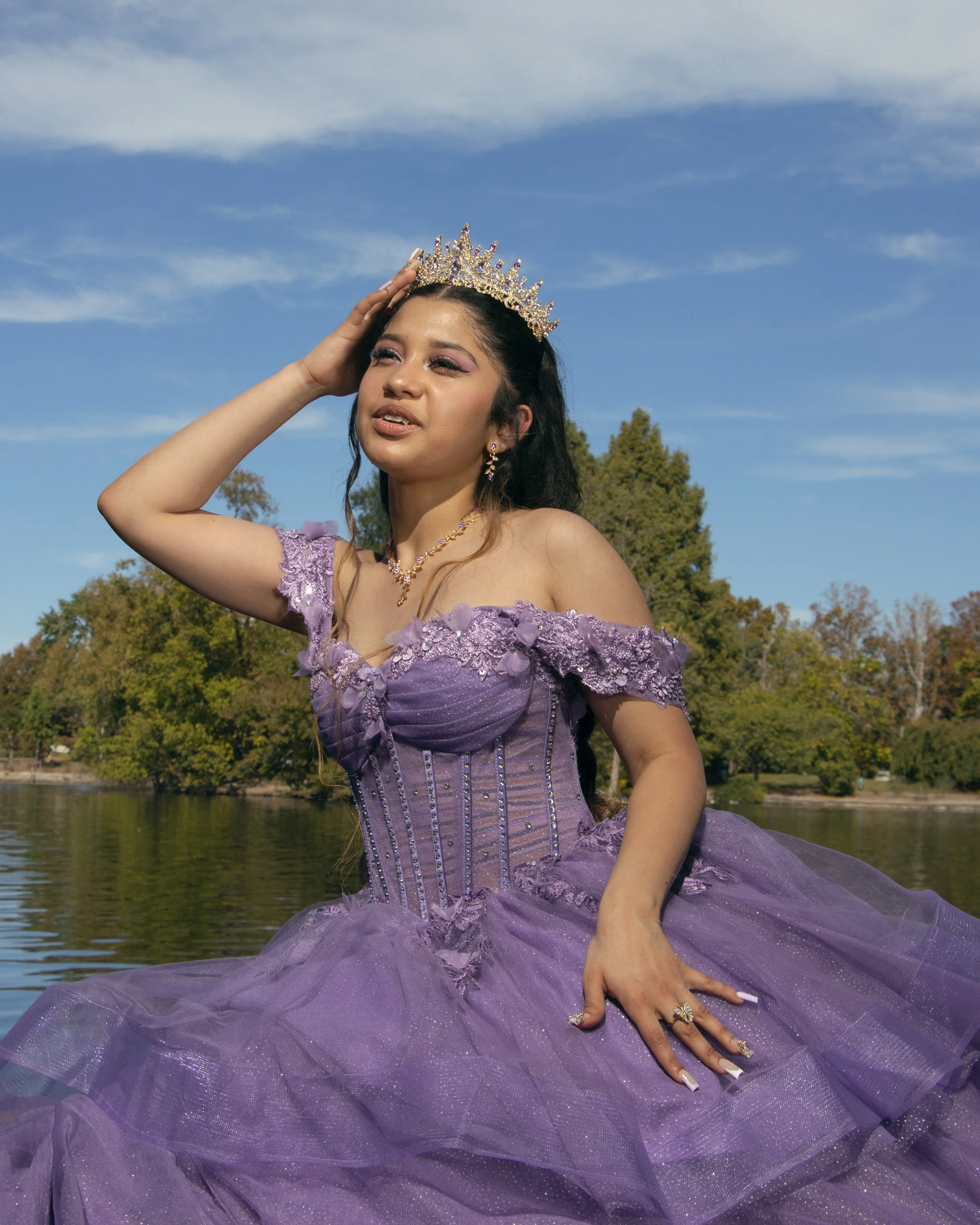 Young woman in a purple ball gown with floral details, wearing a sparkling tiara, posing outdoors by a body of water with trees and a blue sky in the background.